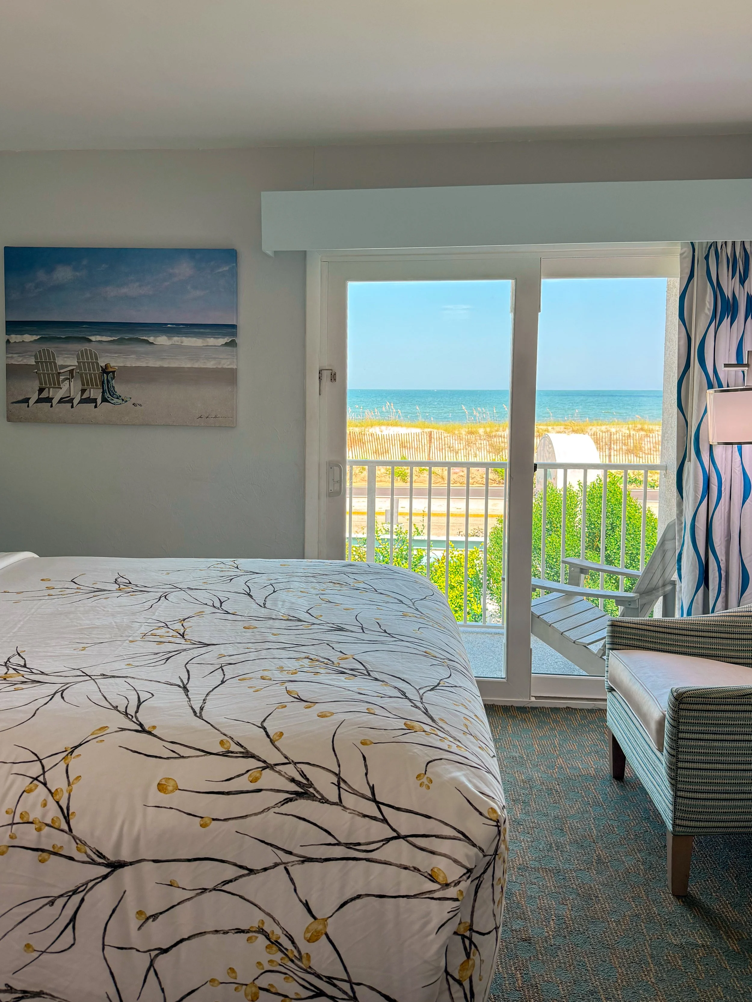 A hotel room with a view of the beach and ocean through a glass door. There is a bed with a patterned bedspread, a white chair, and a painting of a beach scene on the wall.