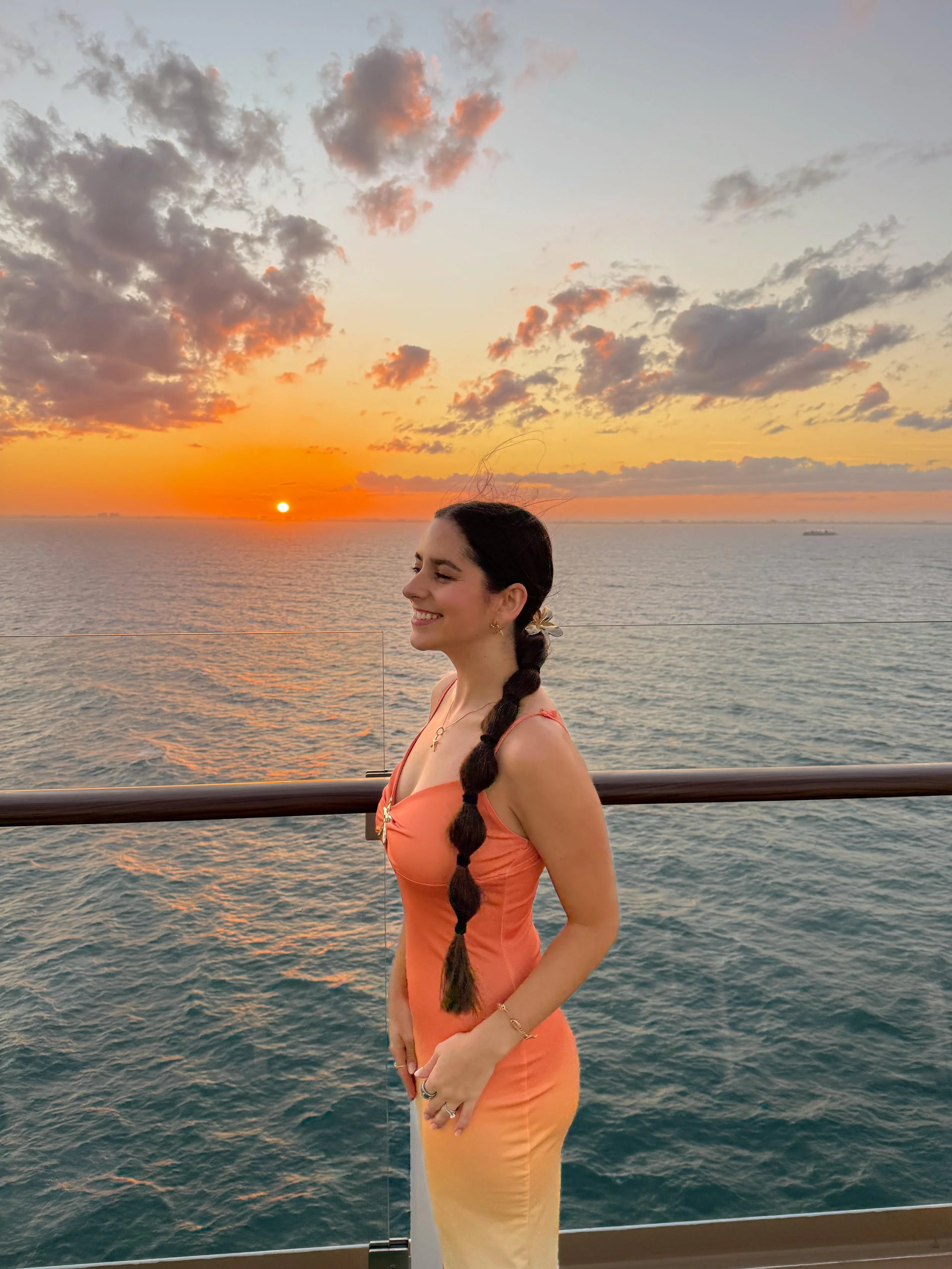 A woman with long dark hair in a braid, wearing an orange dress, is smiling with eyes closed while standing on a ship railing during sunset over the ocean with clouds in the sky.