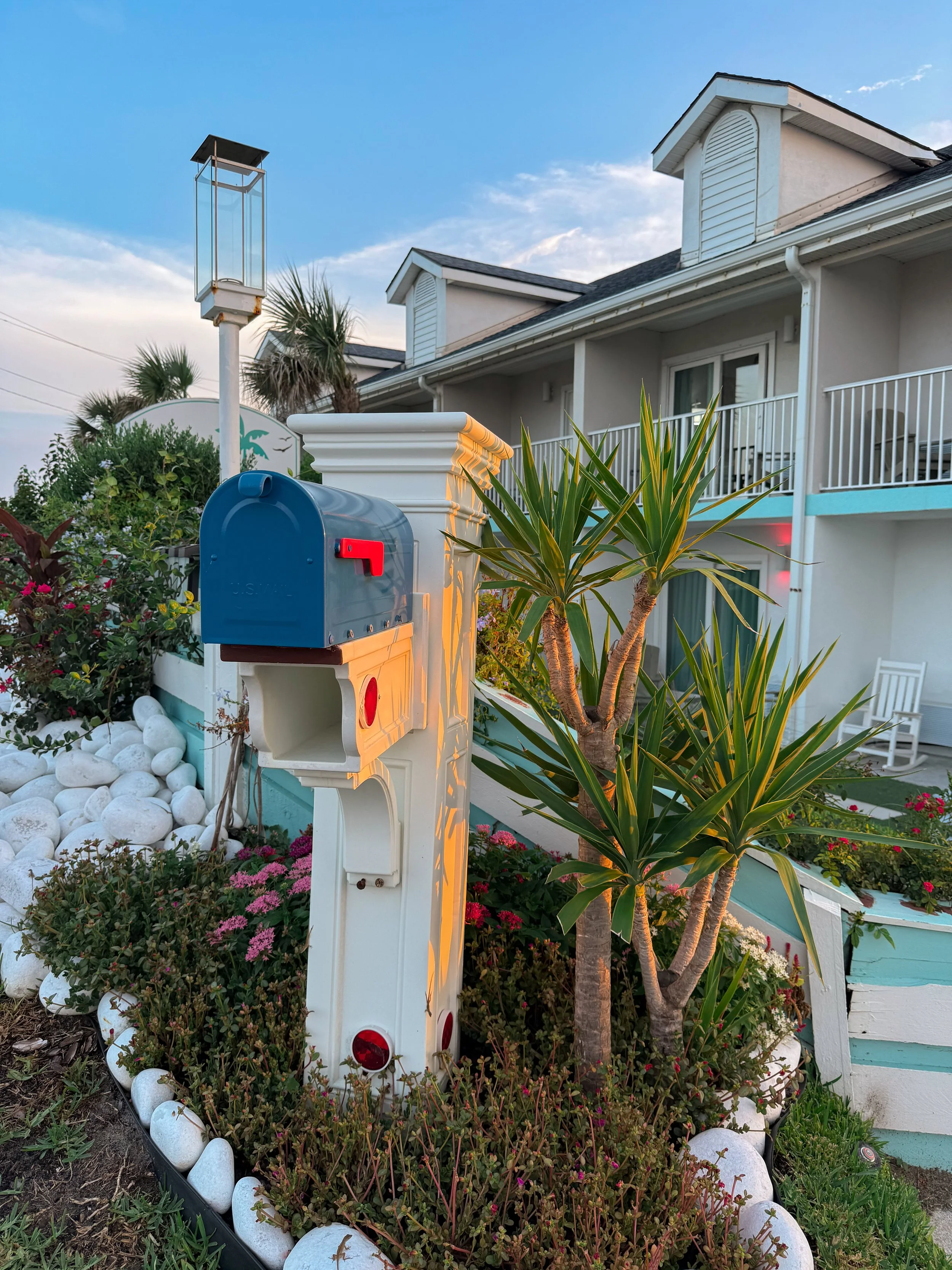 A residential building with a balcony, two dormer windows, and a white exterior. In the flower bed outside, there are green plants, pink flowers, white stones, a blue mailbox, and a tree. There is also a tall, empty lamp post in the foreground.