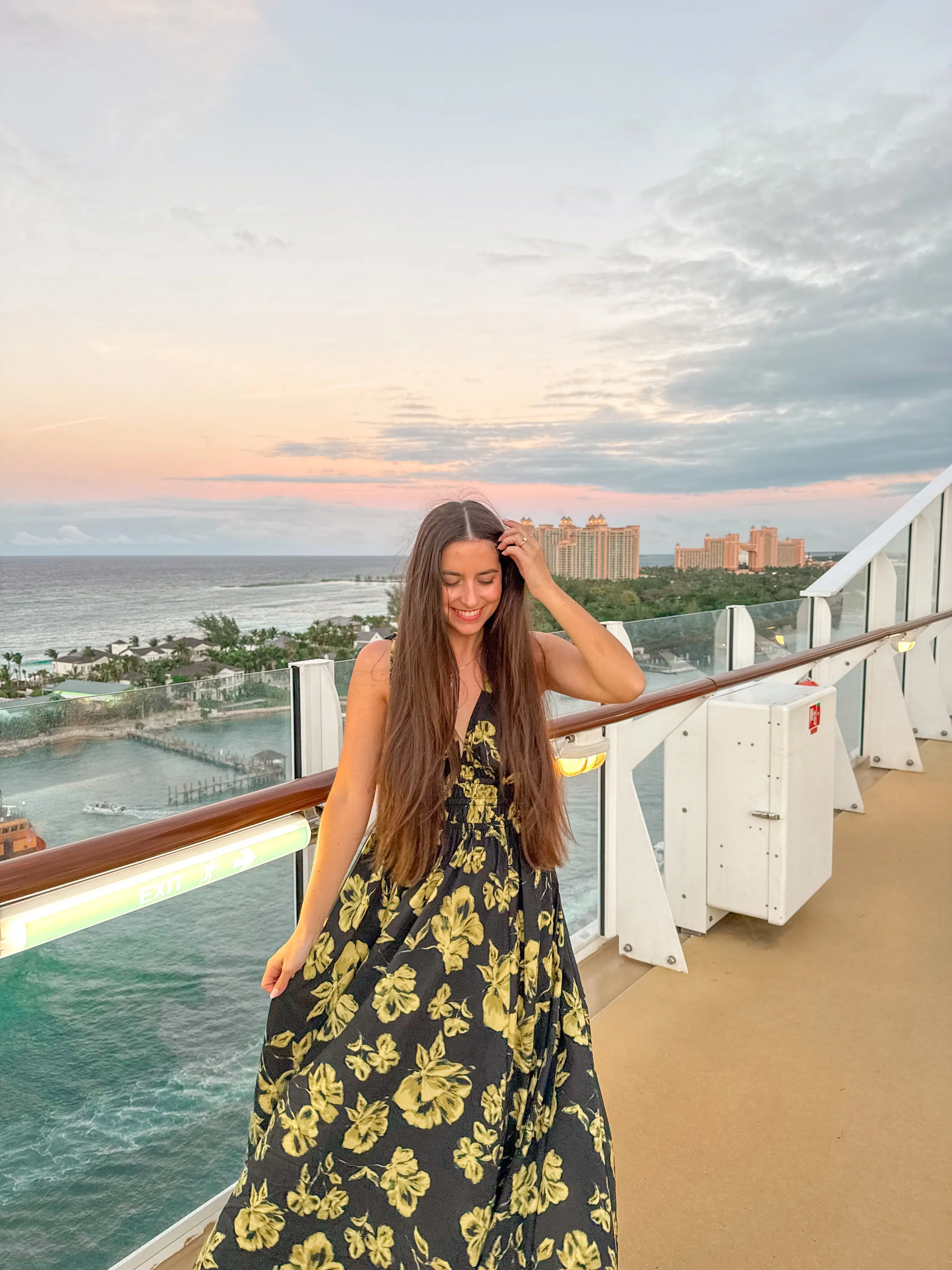 A young woman with long brown hair wearing a black dress with yellow floral pattern, smiling and touching her hair, on a cruise ship balcony overlooking the ocean and a city skyline at sunset.