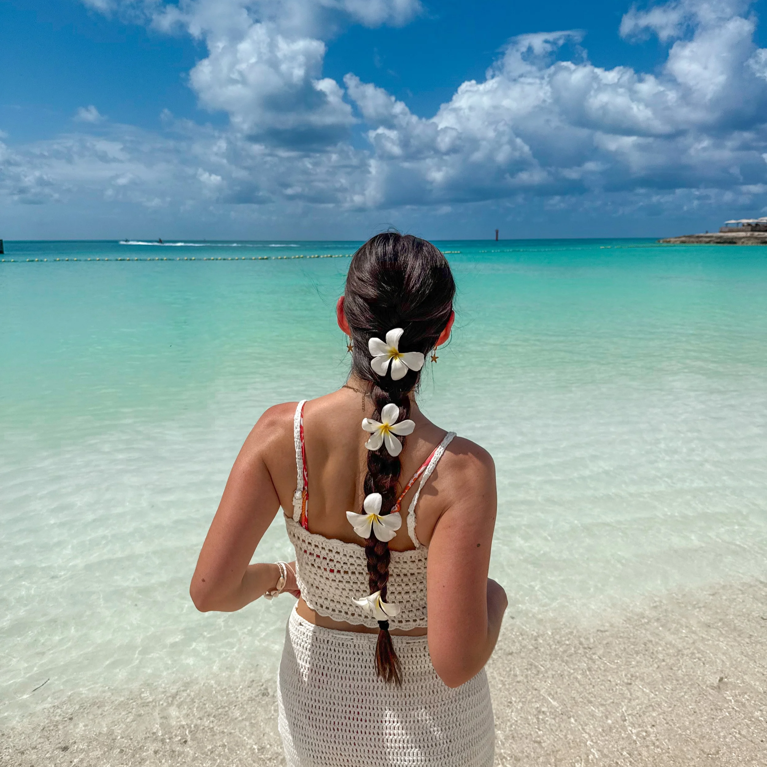 A woman with a long braid decorated with two white flowers stands on a beach facing the ocean. The sky is partly cloudy with blue sky visible, and the water is a turquoise color. There are some structures and a lighthouse or tower in the distance.