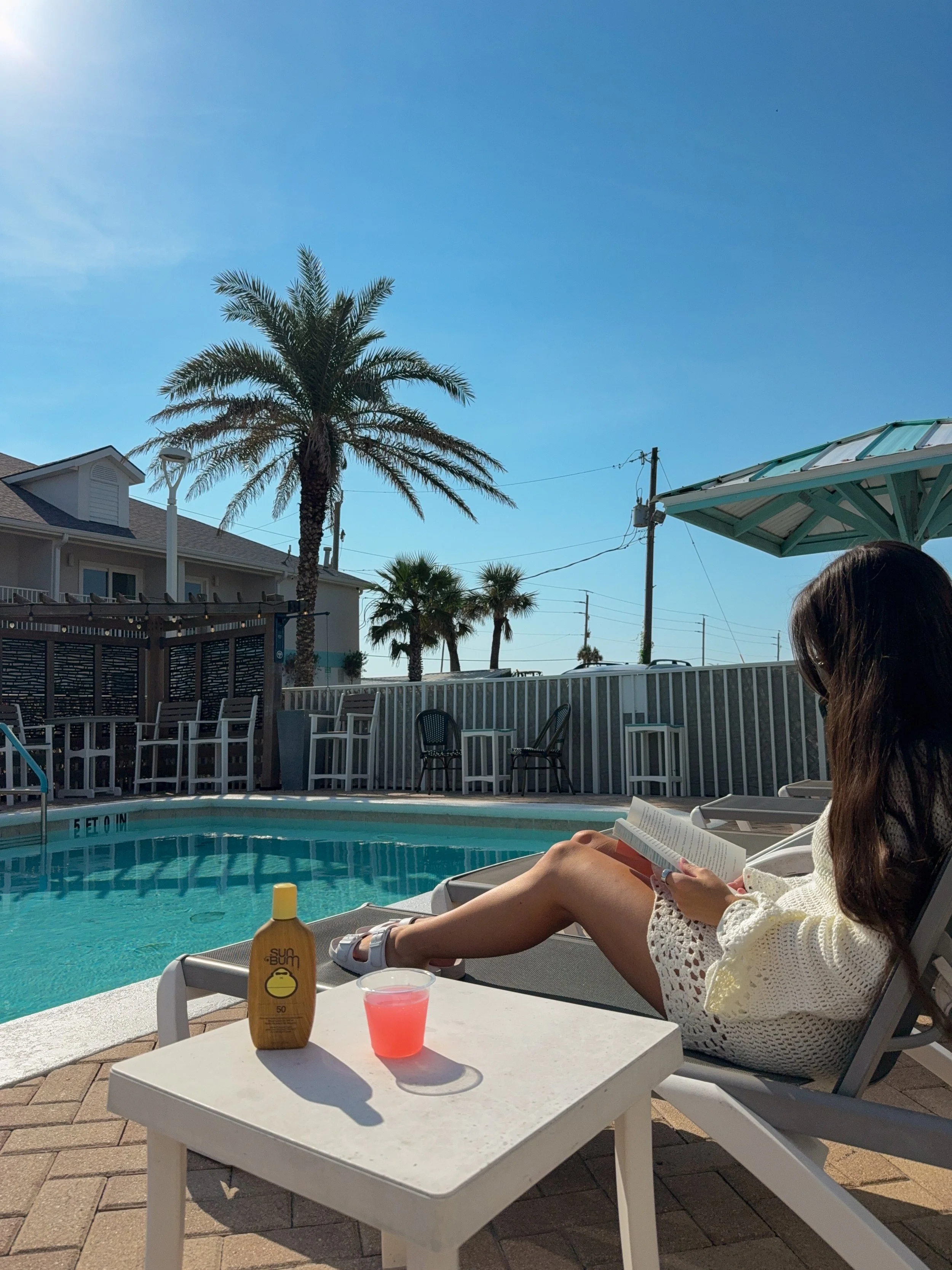A woman sitting by a swimming pool, reading a book, with sunscreen bottle, pink drink, and pool chairs nearby, under a sunny sky.