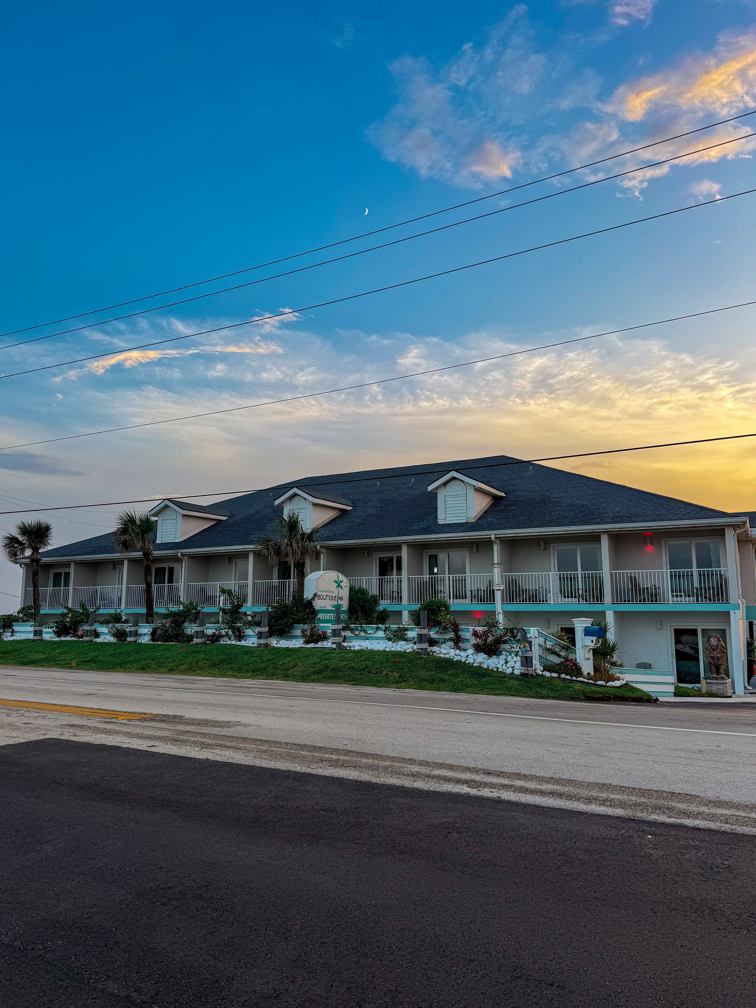 A two-story motel or hotel building with white exterior walls and gray roofing, adorned with palm trees and landscaping in front. The sky above is partly cloudy with a crescent moon visible during sunset or sunrise.