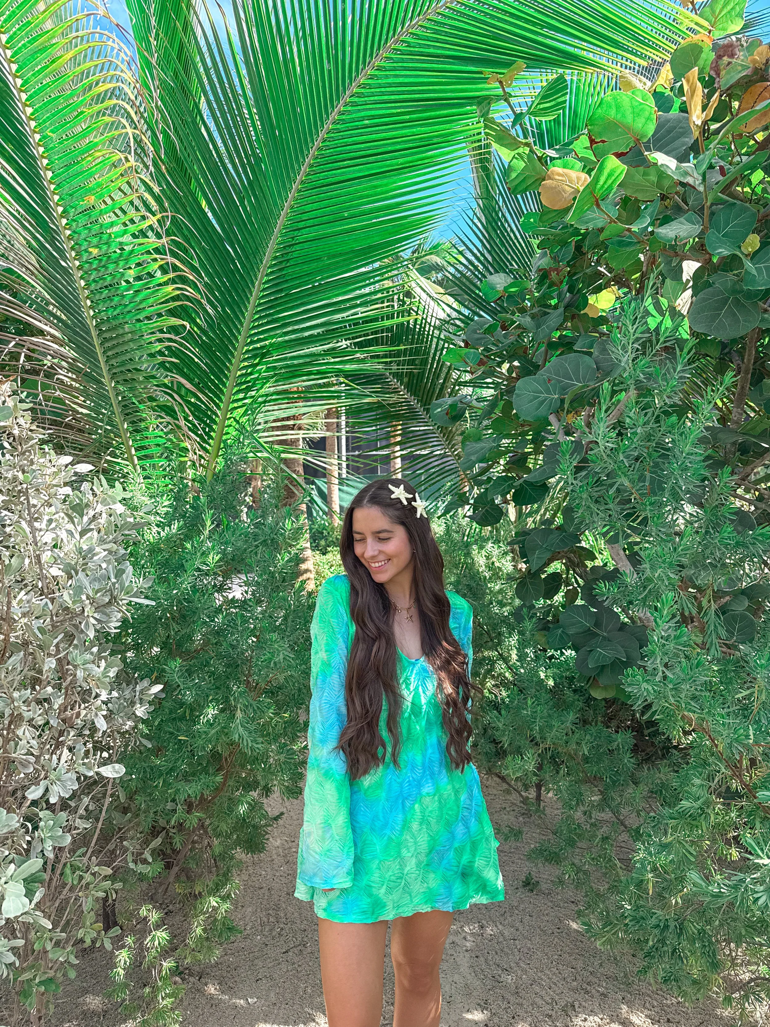 A woman with long dark hair wearing a blue and green patterned dress standing among lush tropical plants with large green and dark leaves, smiling with her eyes closed.