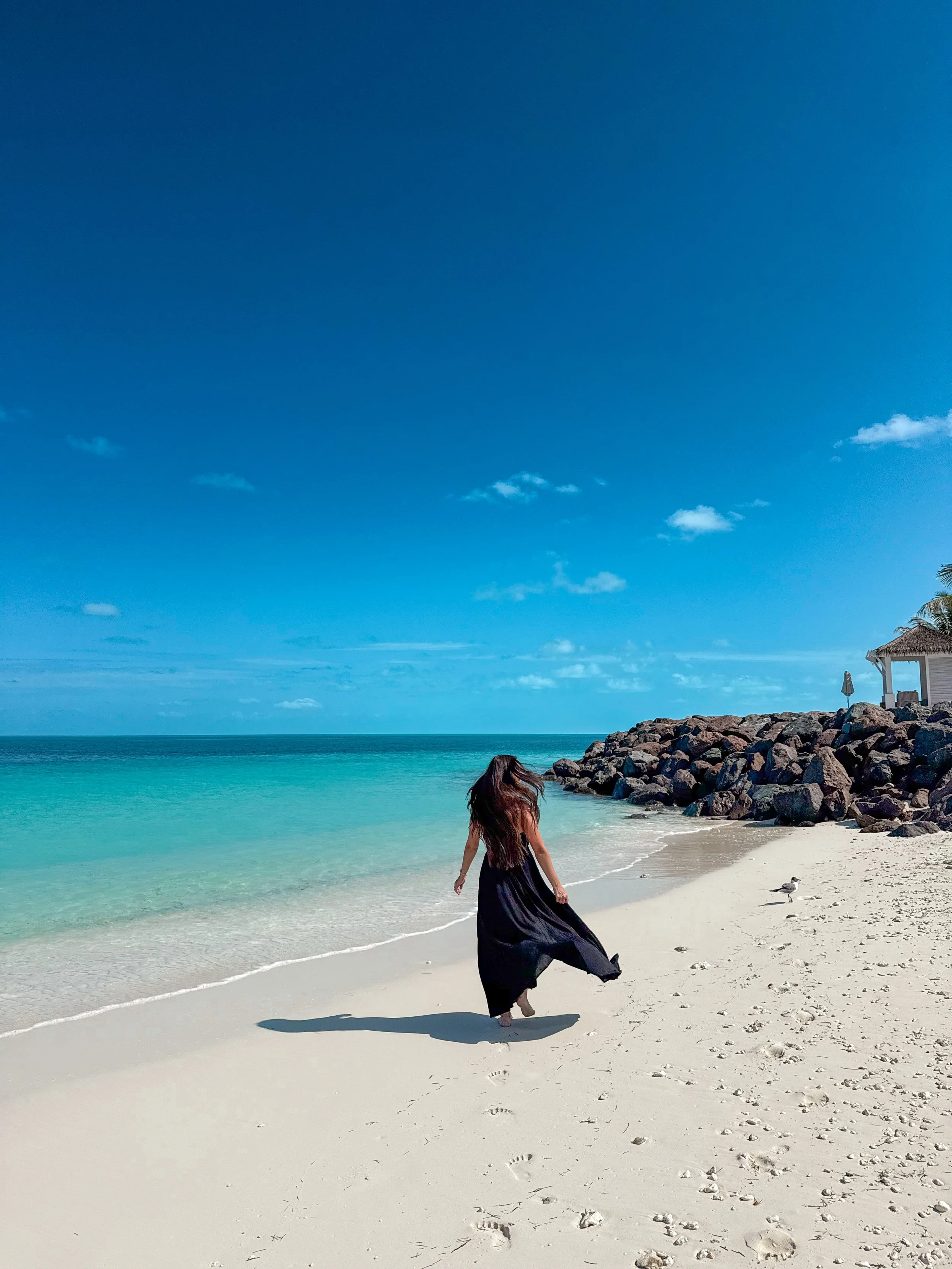 A woman in a long black dress walking along a sandy beach near turquoise water and rocks, with a blue sky overhead and a small thatched shelter on the right side of the image.