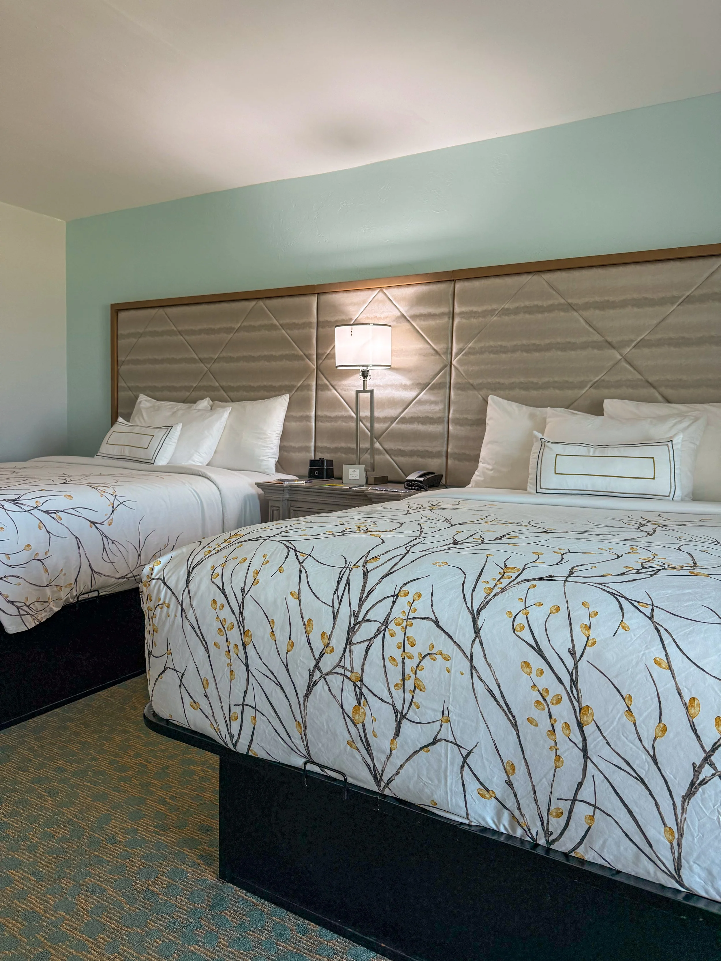 Two large hotel beds with white bedding and decorative pillows, a bedside table with a lamp, phone, and notepad, in a hotel room with a beige and light blue color scheme.