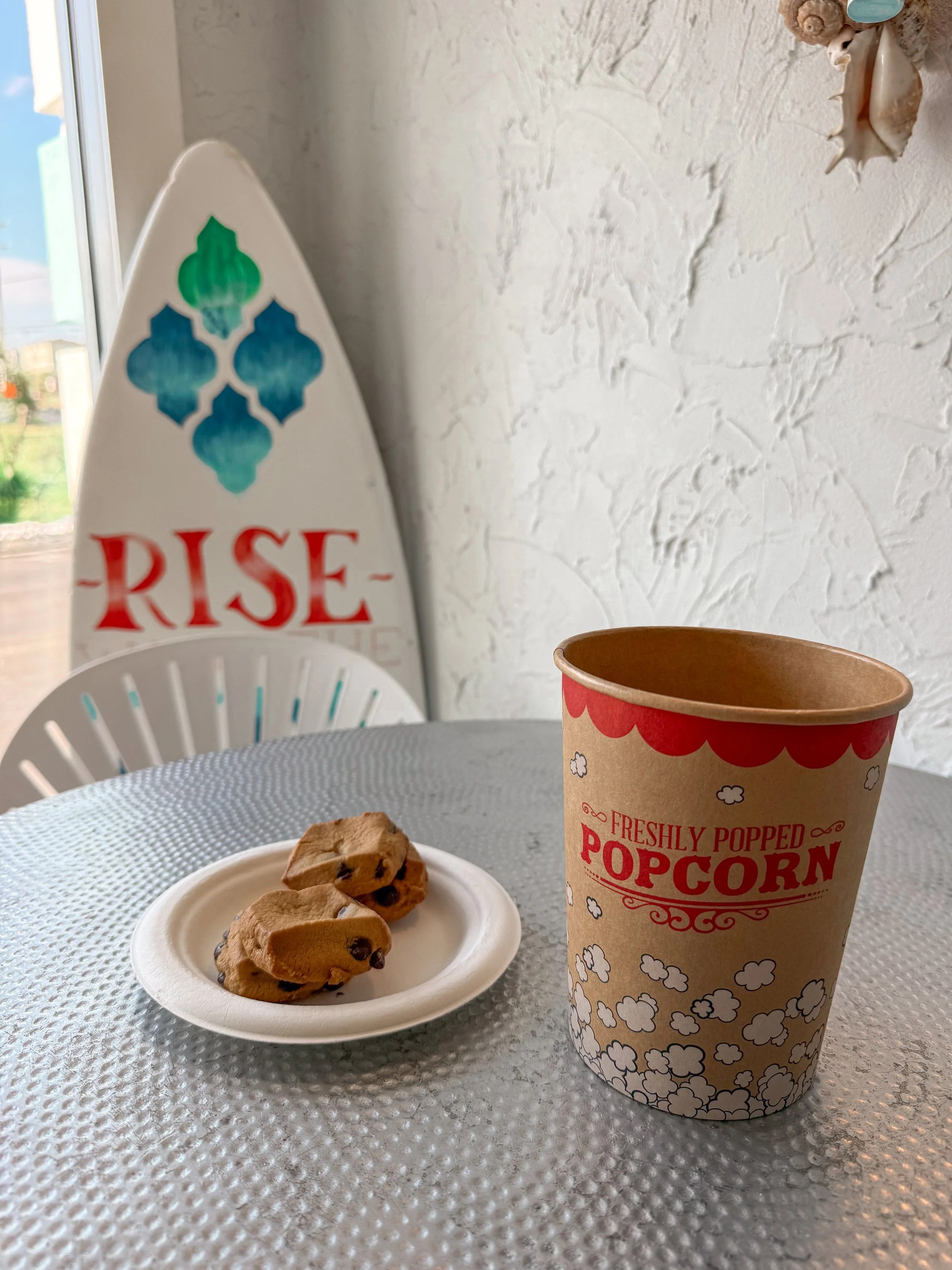 A paper cup of popcorn, a small white plate with chocolate chip cookies, and a cookie sheet with chocolate chip cookies on a textured white table with a white textured wall in the background.