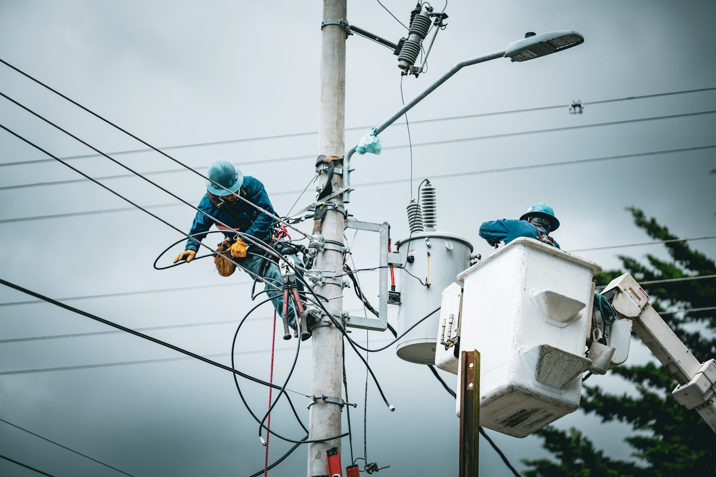 Two utility workers wearing helmets and safety gear are working on an electric power pole, using a bucket truck to reach the high electrical components and wires on a cloudy day.