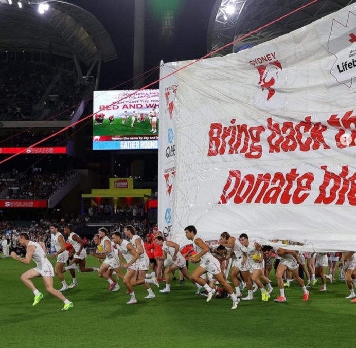 Australian football team the Sydney Swans runs onto the field in white jerseys. They're breaking through a large banner that reads 'Bring back the red. Donate blood.'