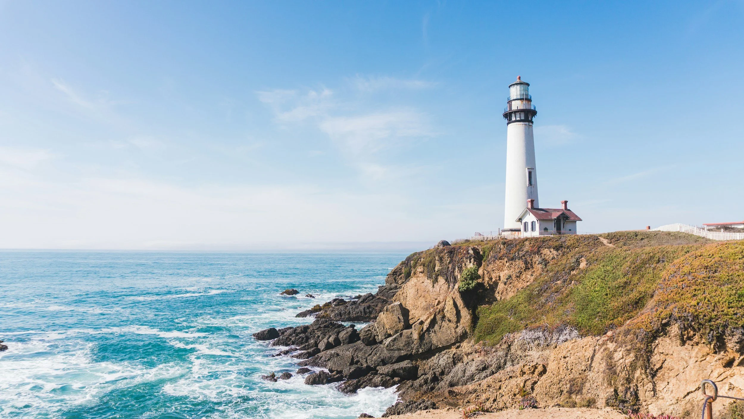 A lighthouse stands on a rocky cliff overlooking the ocean under a clear blue sky.