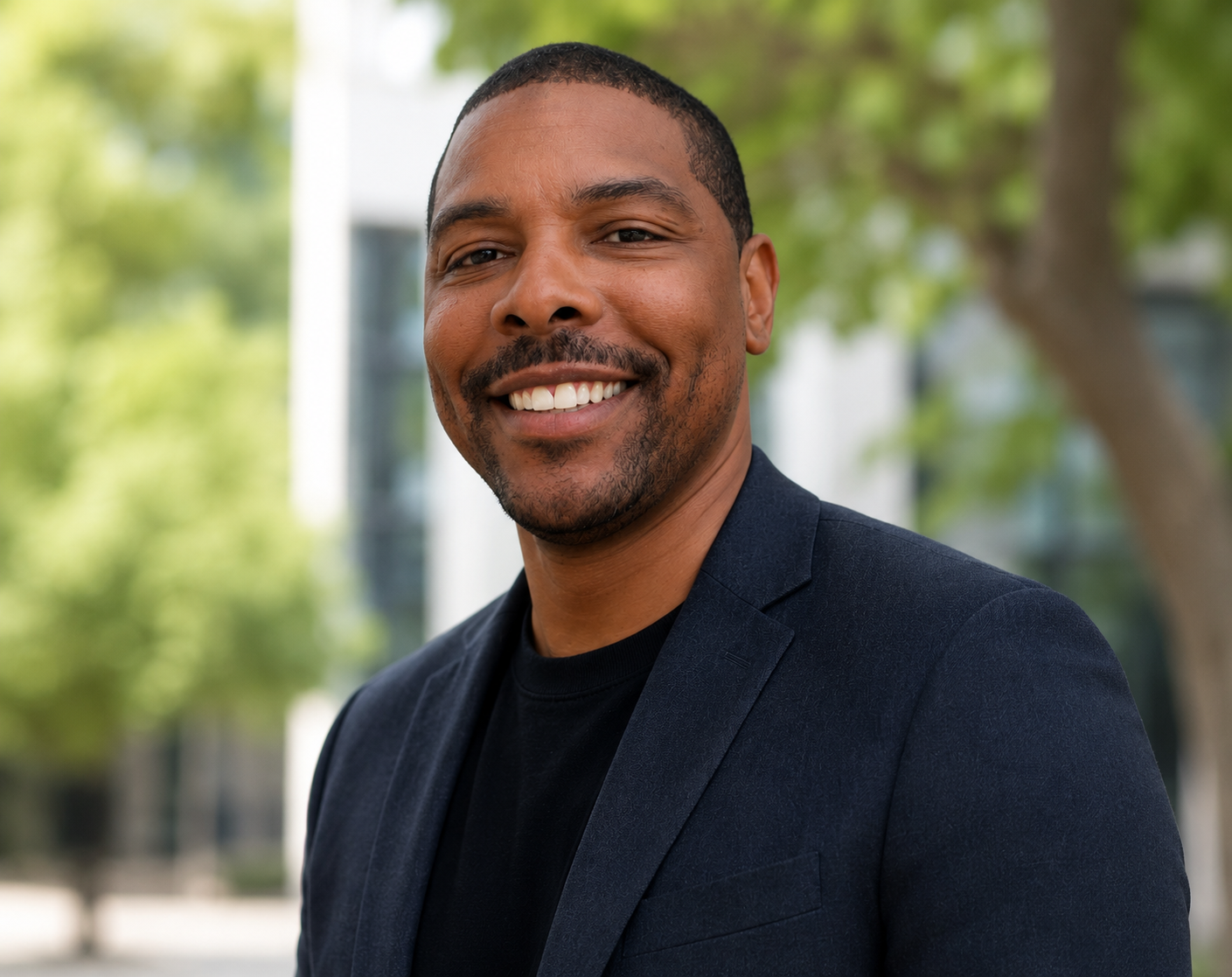 Portrait of a smiling young man with a beard, wearing a black shirt and dark blazer, outdoors with green trees and building in the background.