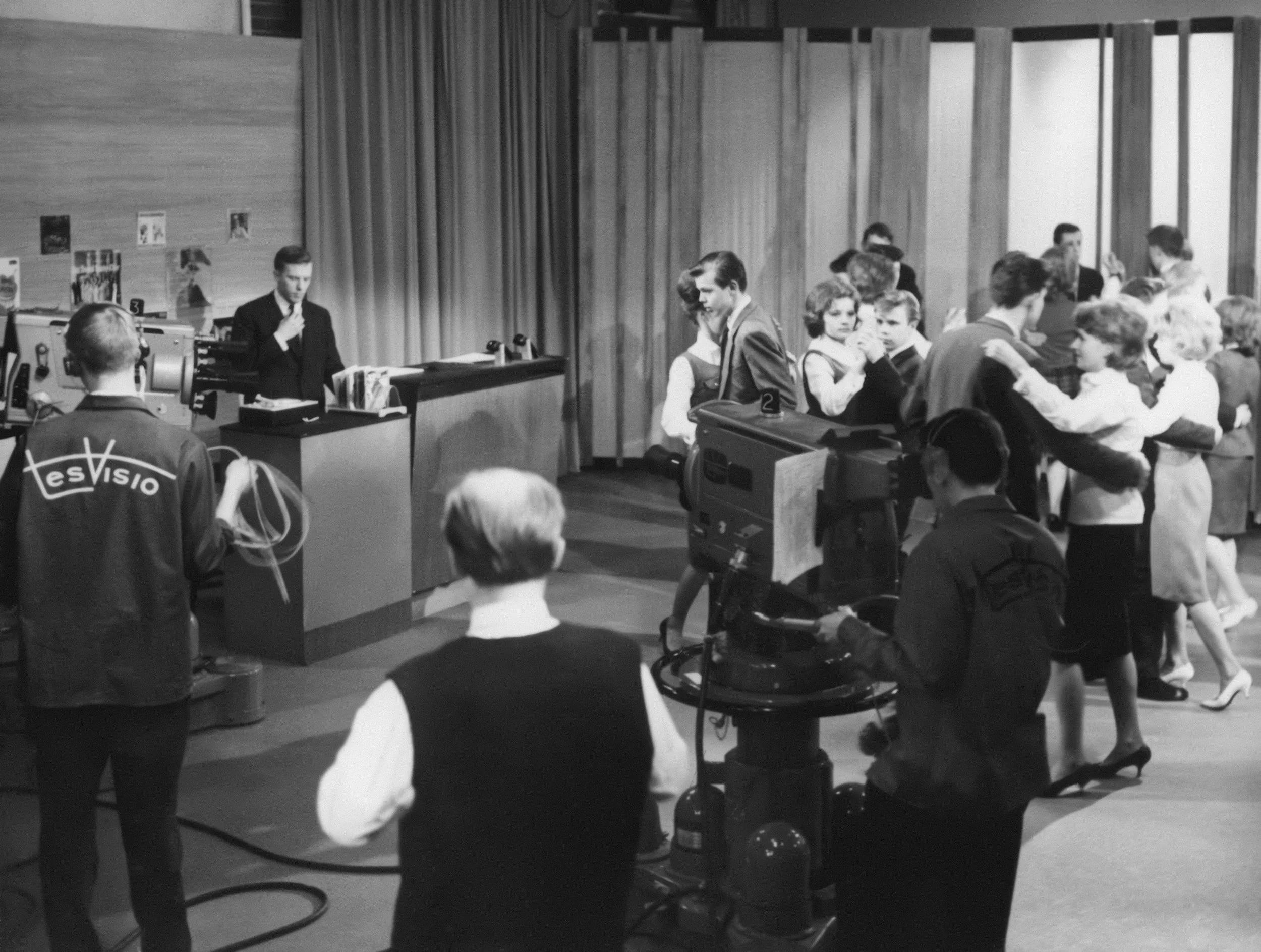 A black and white photo of a busy reception area with people waiting and interacting. Some individuals appear to be holding or carrying luggage, suggesting this might be an airport or train station check-in or waiting area. There are desks, a curtain, and several people engaged in various activities, with some facing away from the camera and others speaking to staff or each other.