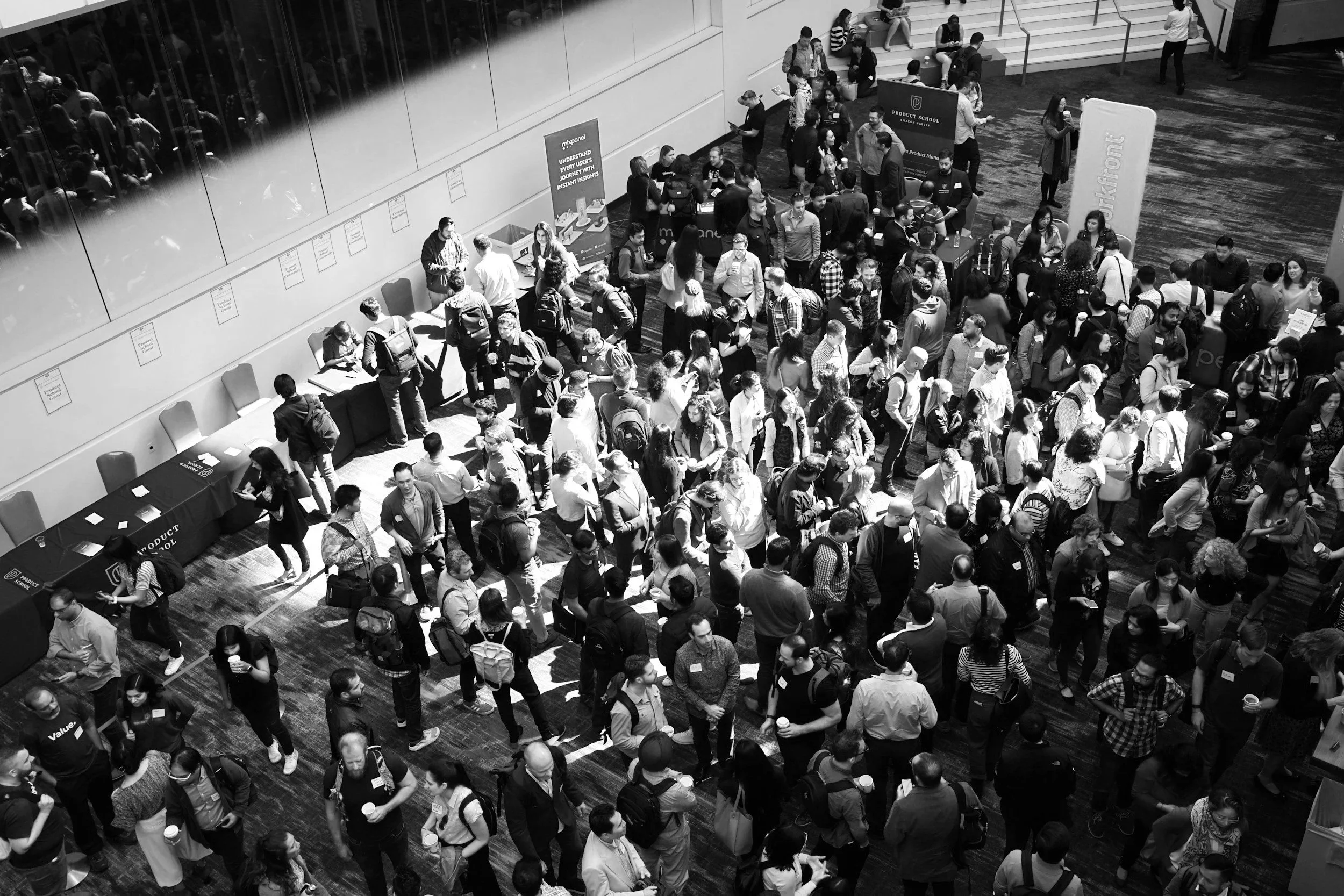 A large crowd of people attending a conference or event in a spacious hall, with many sitting and some standing, engaging with booths and displays.