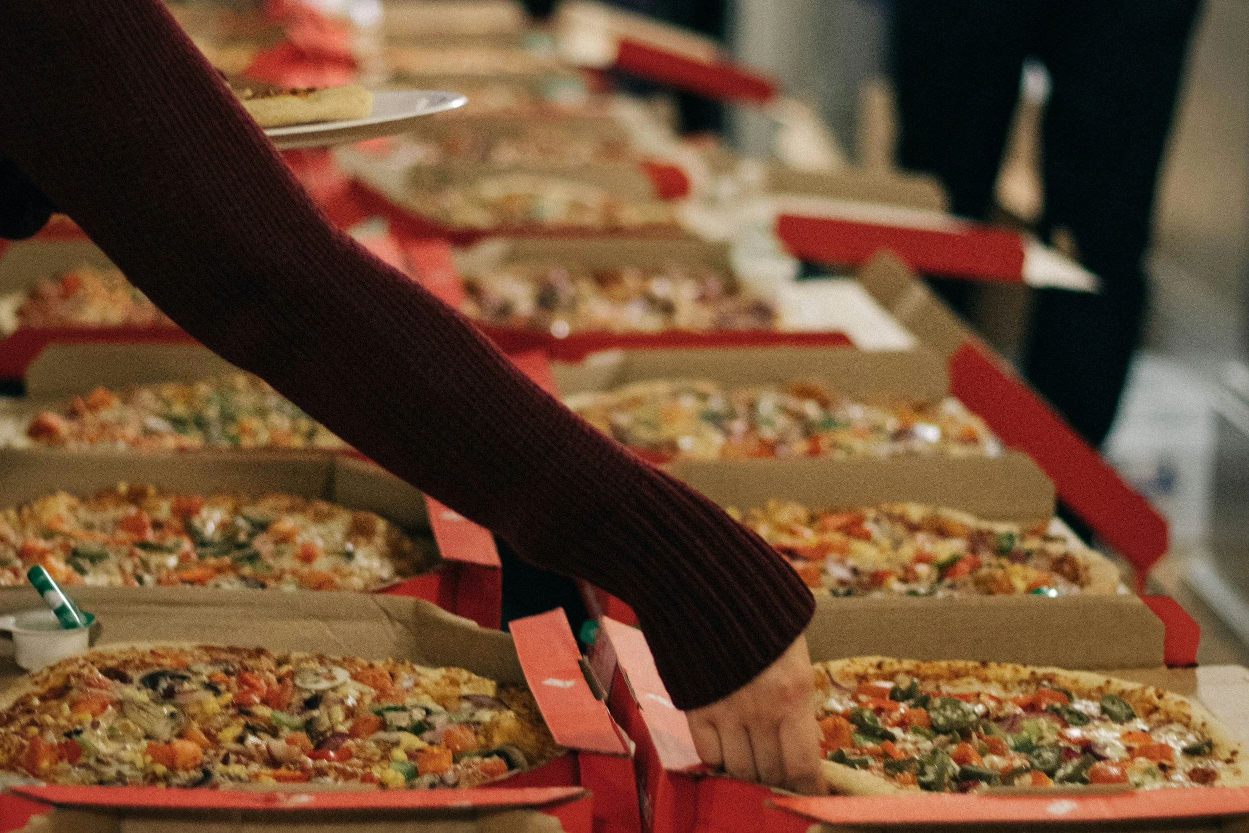 Multiple pizza boxes with various pizzas displayed on a table. Someone is reaching for a slice of pizza from one of the boxes.