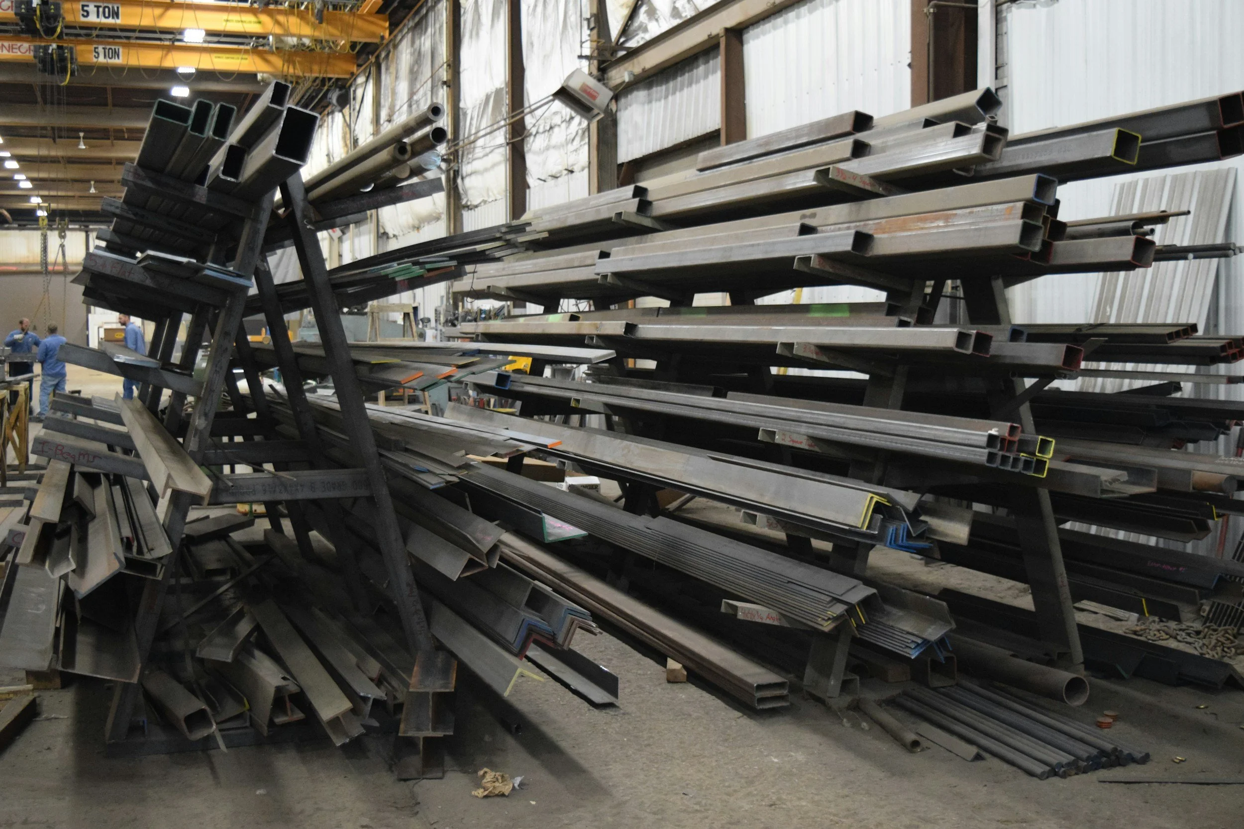 Stacks of metal rectangular tubing stored on racks inside a warehouse or industrial facility, with workers visible in the background.