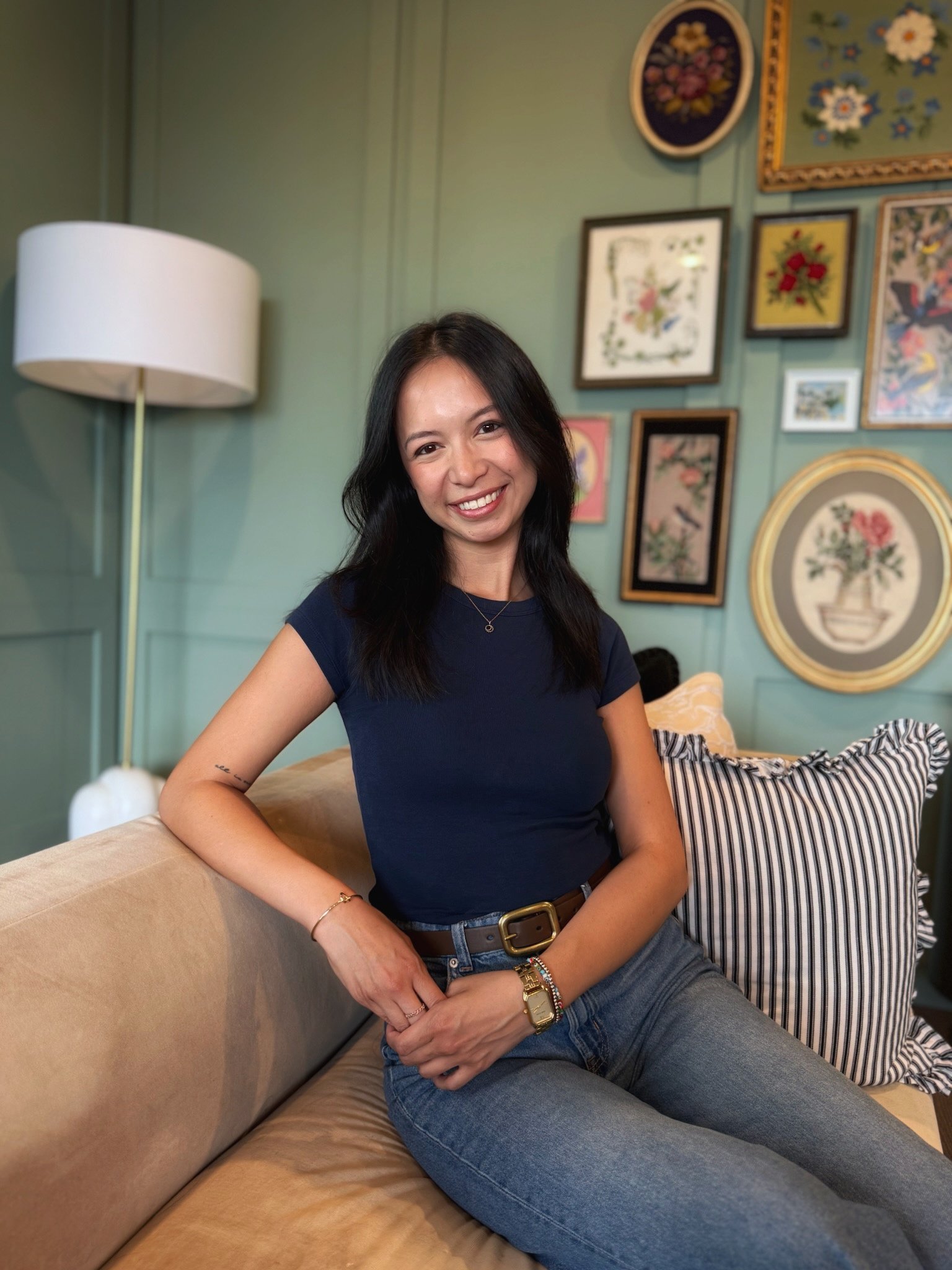 A woman with long black hair, wearing a dark blue top and jeans, sitting on a beige sofa with a striped pillow, smiling at the camera, in a room with green walls decorated with framed floral artwork and a white floor lamp.