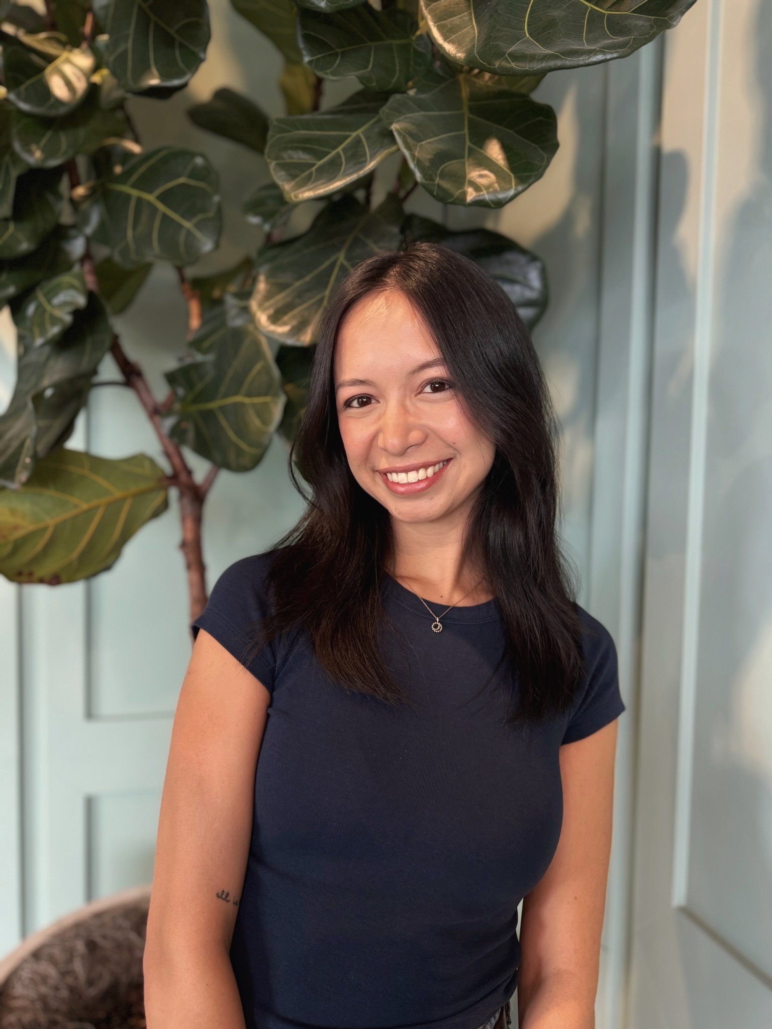 A woman with long black hair smiling in front of a large green plant, wearing a navy blue t-shirt and a necklace with a small pendant, indoors.