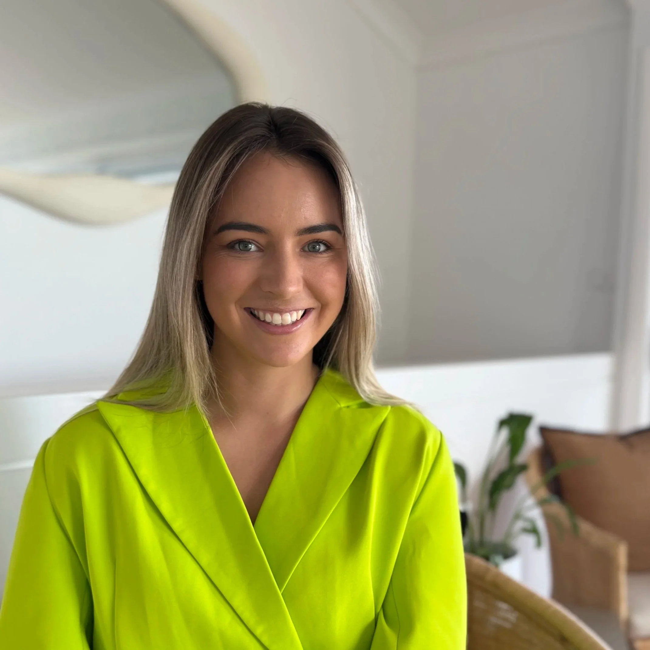 A woman with long blonde hair and blue eyes is smiling, wearing a bright yellow-green safety jacket, standing indoors with a blurred background of a white wall and some plants in Newcastle Australia
