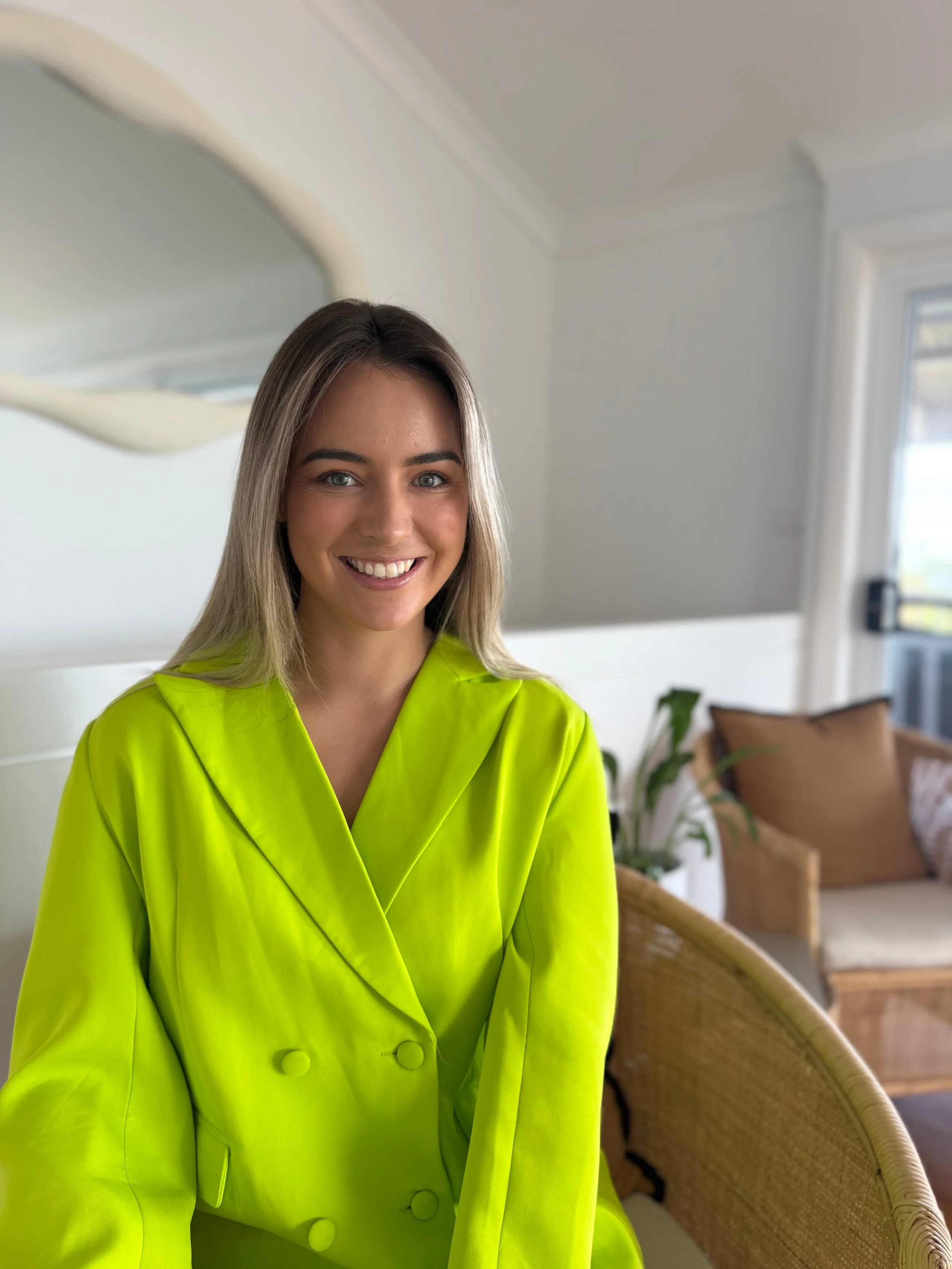 A young woman with blonde hair and blue eyes smiling, wearing a bright yellow blazer, sitting on a wicker chair in a well-lit room with a window and a plant in the background in Newcastle Australia
