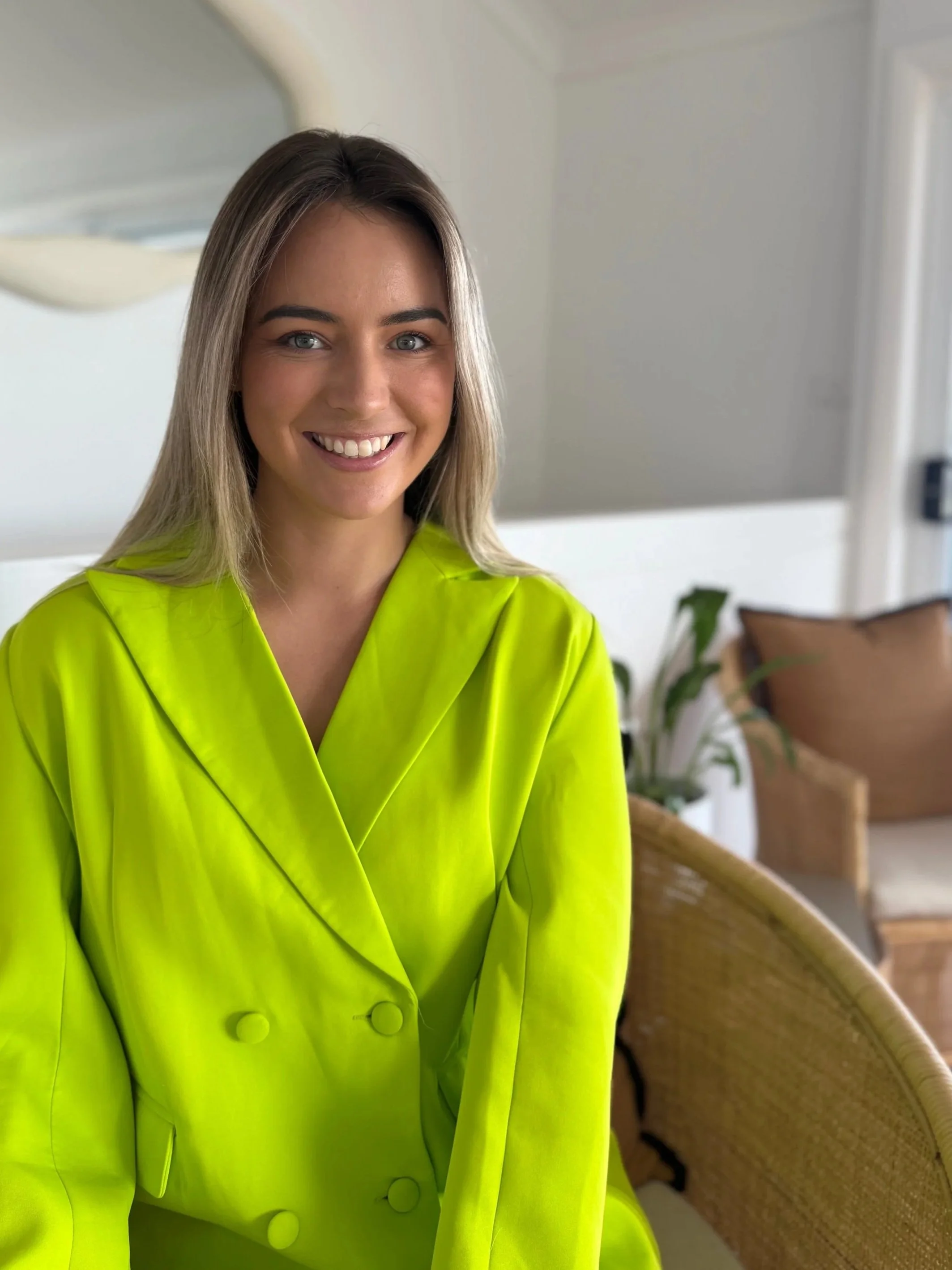 A finance broker with blonde hair and blue eyes smiling, wearing a bright yellow blazer, sitting in a room with white walls in Newcastle Australia
