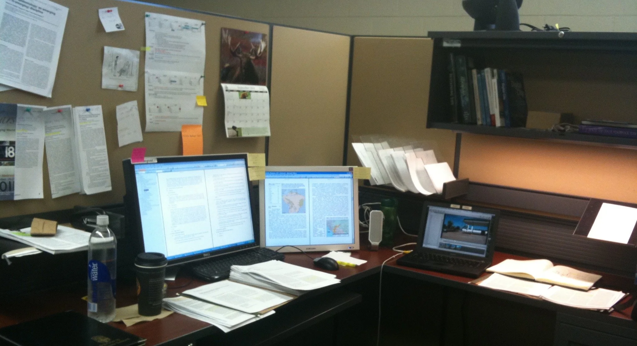 Office desk from 2010, with multiple computer monitors displaying documents and maps, papers and notebooks scattered across the desk, bulletin board with notices and posters in the background.