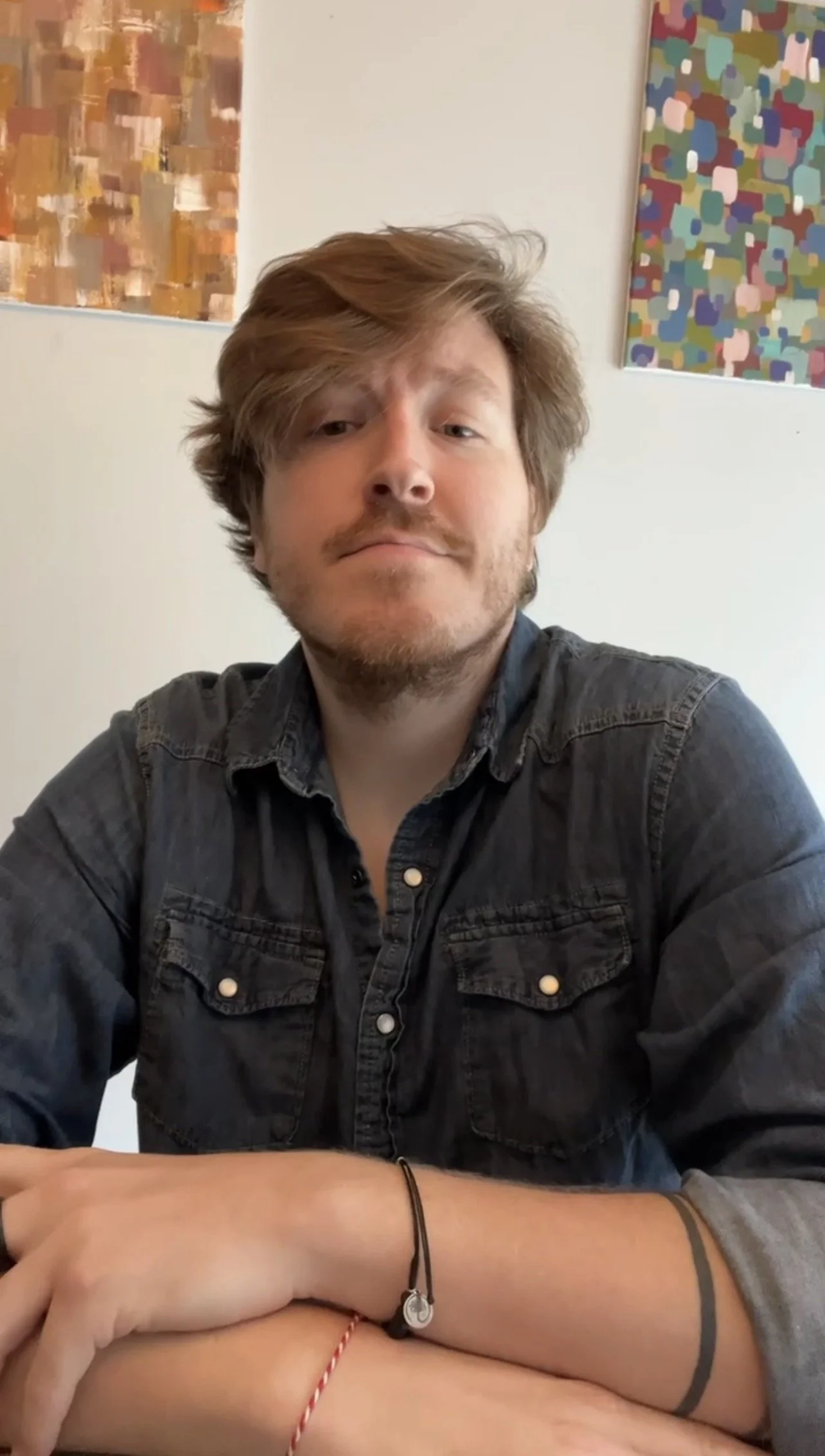 A man with light brown, messy hair and a beard wearing a dark button-up shirt, sitting at a table with art pieces on the wall behind him.
