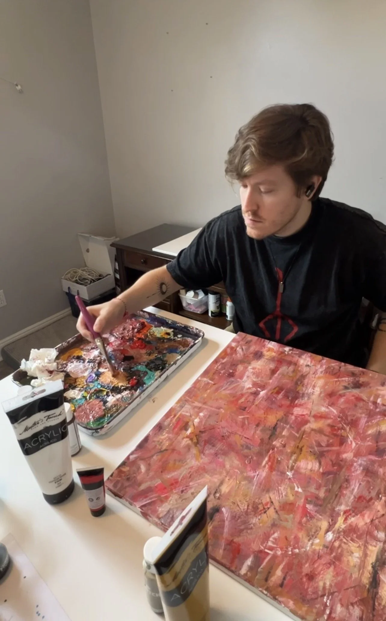 A person with brown hair and a black shirt is sitting at a table, painting with a brush on an abstract painting. The table has various art supplies including paint tubes and a palette containing multiple colors.