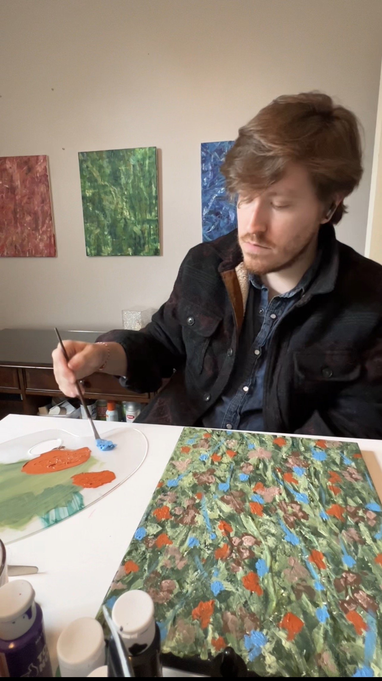 A man is painting on a canvas with a paintbrush in his hand. The painting features a floral or abstract design with bright orange, blue, and green colors. Behind him, there are framed paintings on the wall, and on the table, there are various art supplies and colorful paintings.