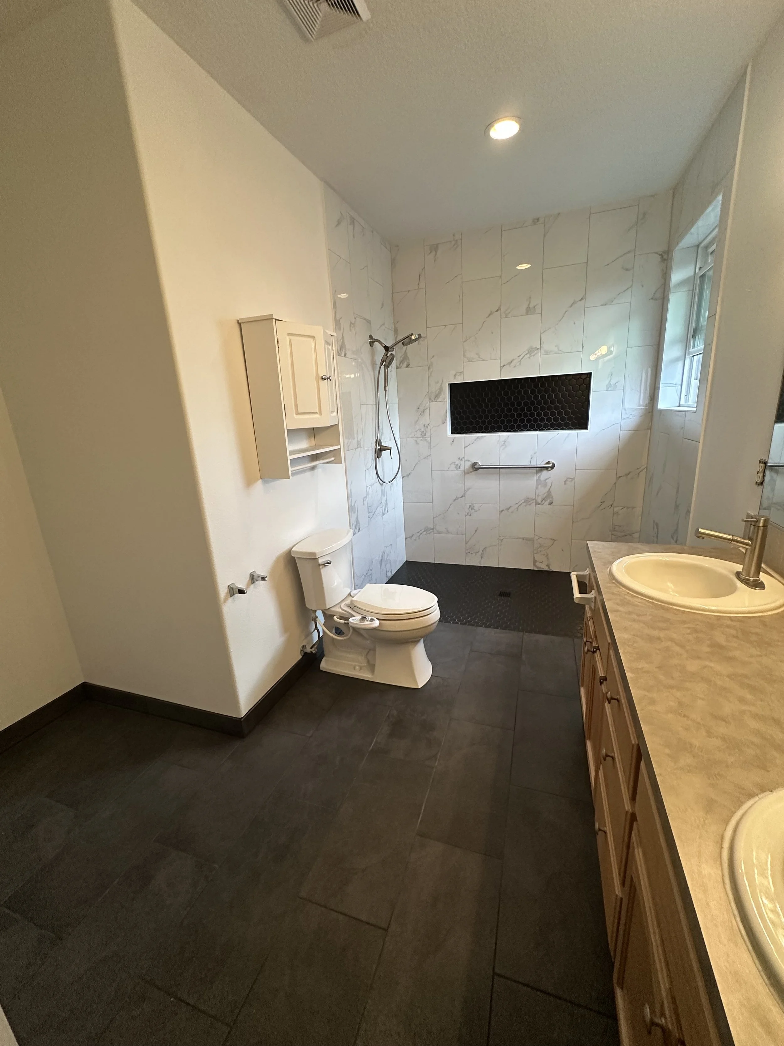 A bathroom with a toilet, a shower area with a black honeycomb shower head on the wall, marble tiles, a window, a small cabinet, and a sink with a beige countertop.