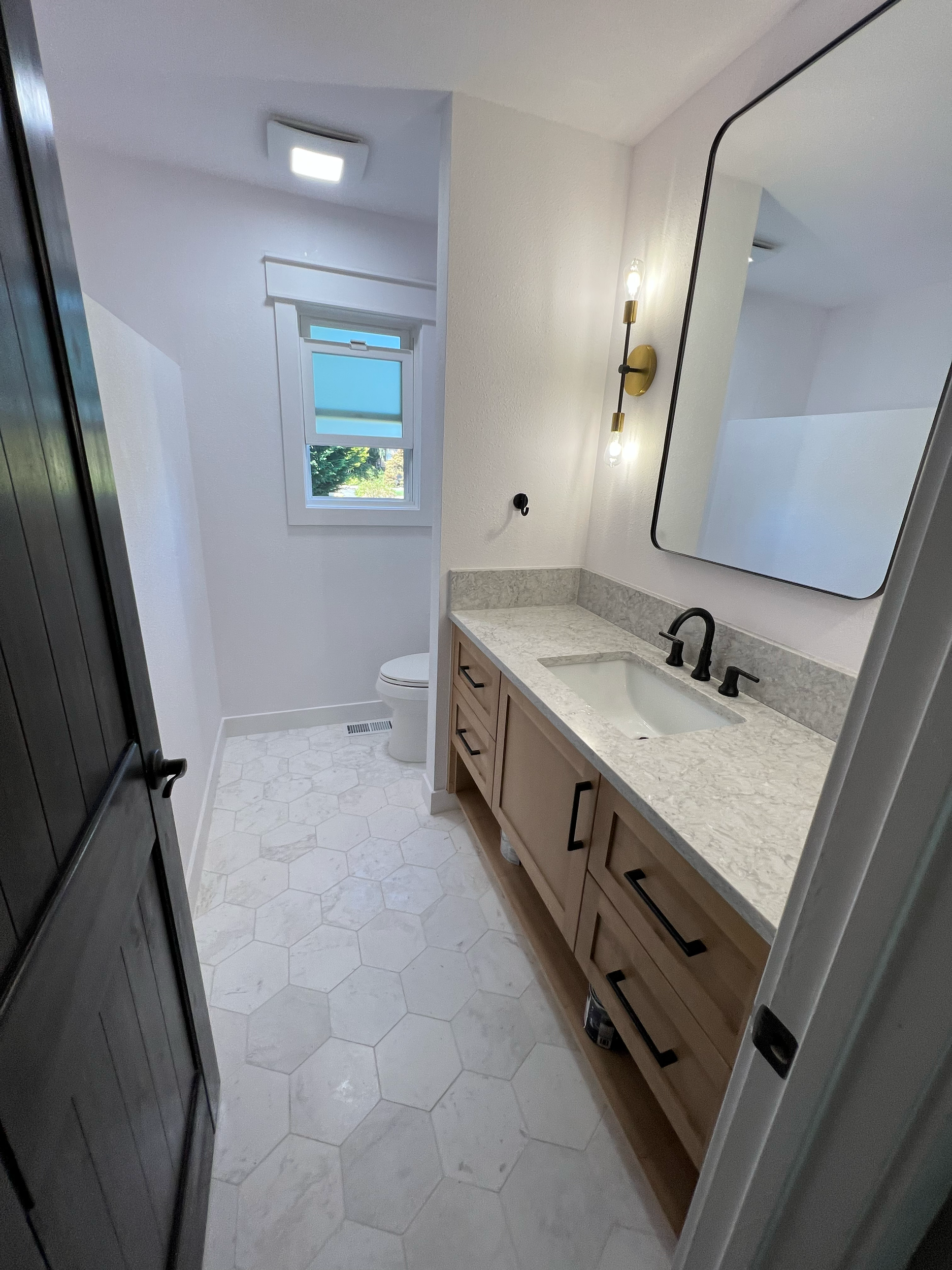 A bathroom with a vanity mirror, a sink with a black faucet, a beige cabinet, a wall sconce with two lights, a window with frosted glass, and part of a shower or closet door on the left.