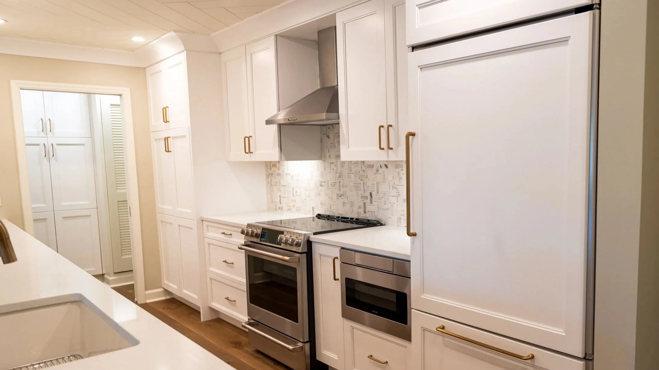 White kitchen with stainless steel stove, oven, range hood, and refrigerator, white cabinets with brass handles, and light-colored walls and ceiling.
