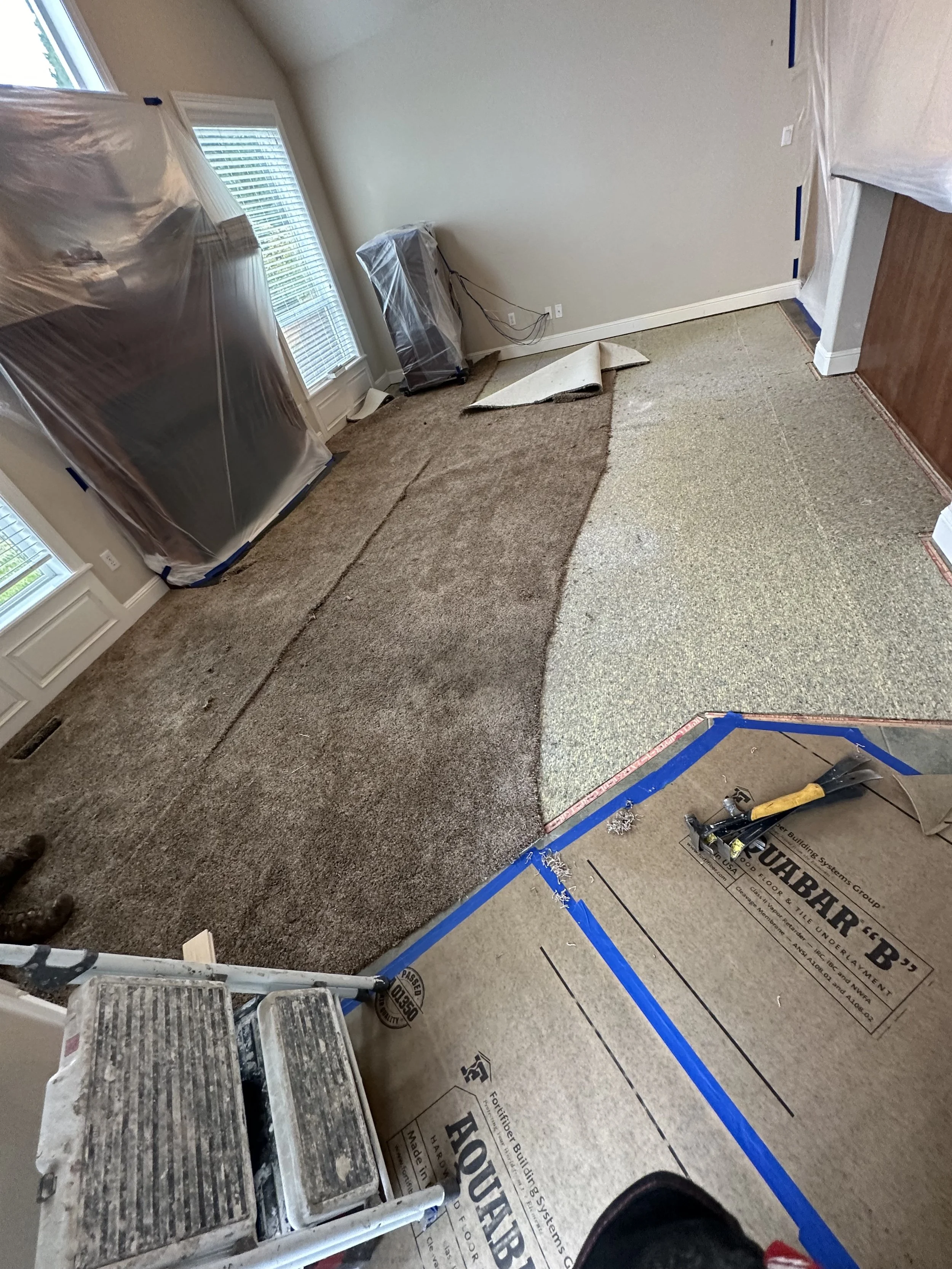 Room under renovation with partially removed flooring, a ladder, covered appliances, and construction tools. The area is taped off with blue painter's tape.
