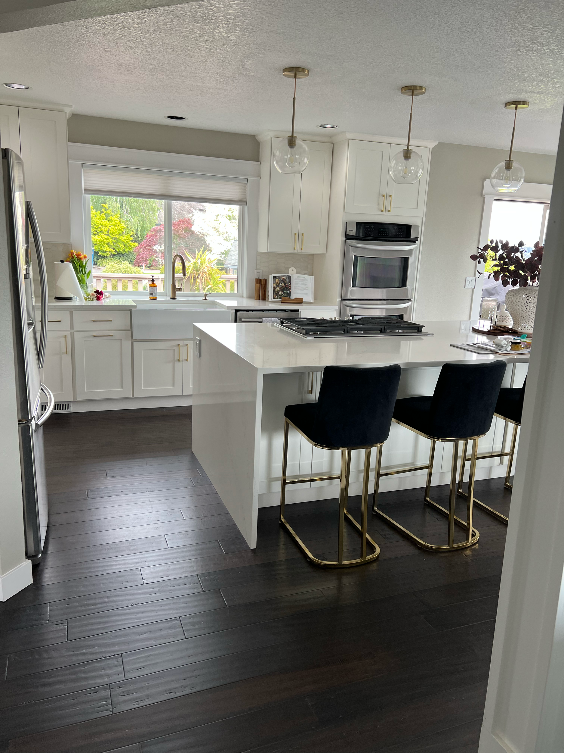 A modern kitchen with white cabinets, dark wood floors, and a white kitchen island with three black barstools with gold legs. Three hanging pendant lights with glass globes are above the island. A window behind the sink shows a view of green trees and plants outside.