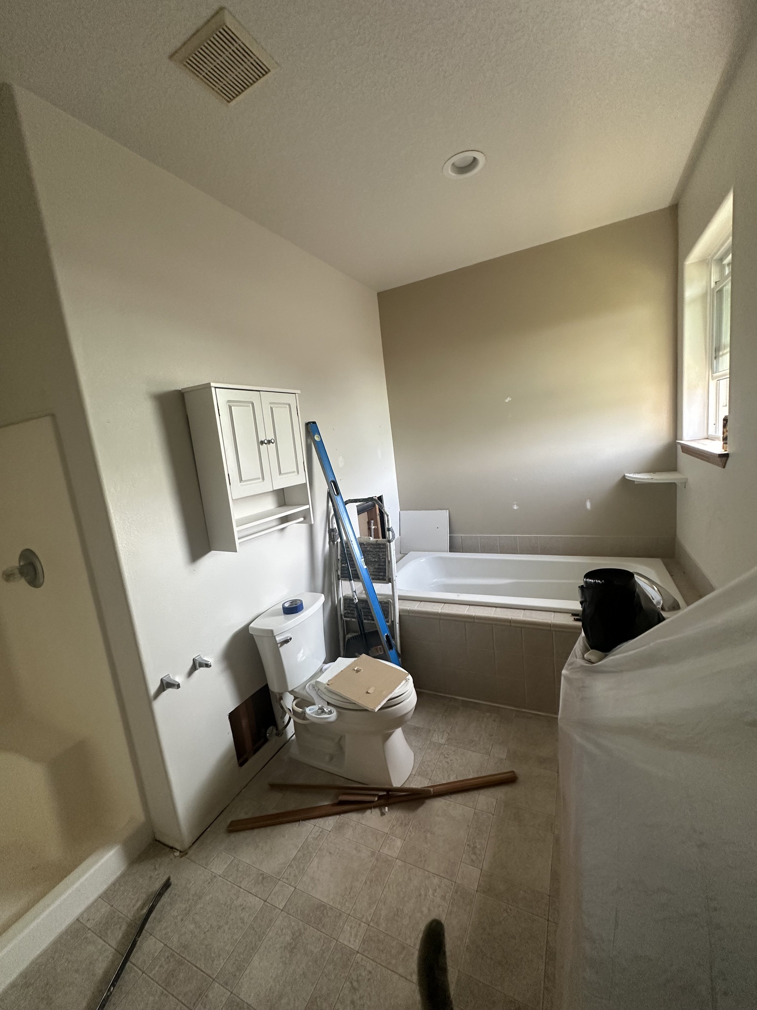 Unfinished bathroom with a toilet, a white wall-mounted cabinet, ladder, and various construction materials and tools scattered around.