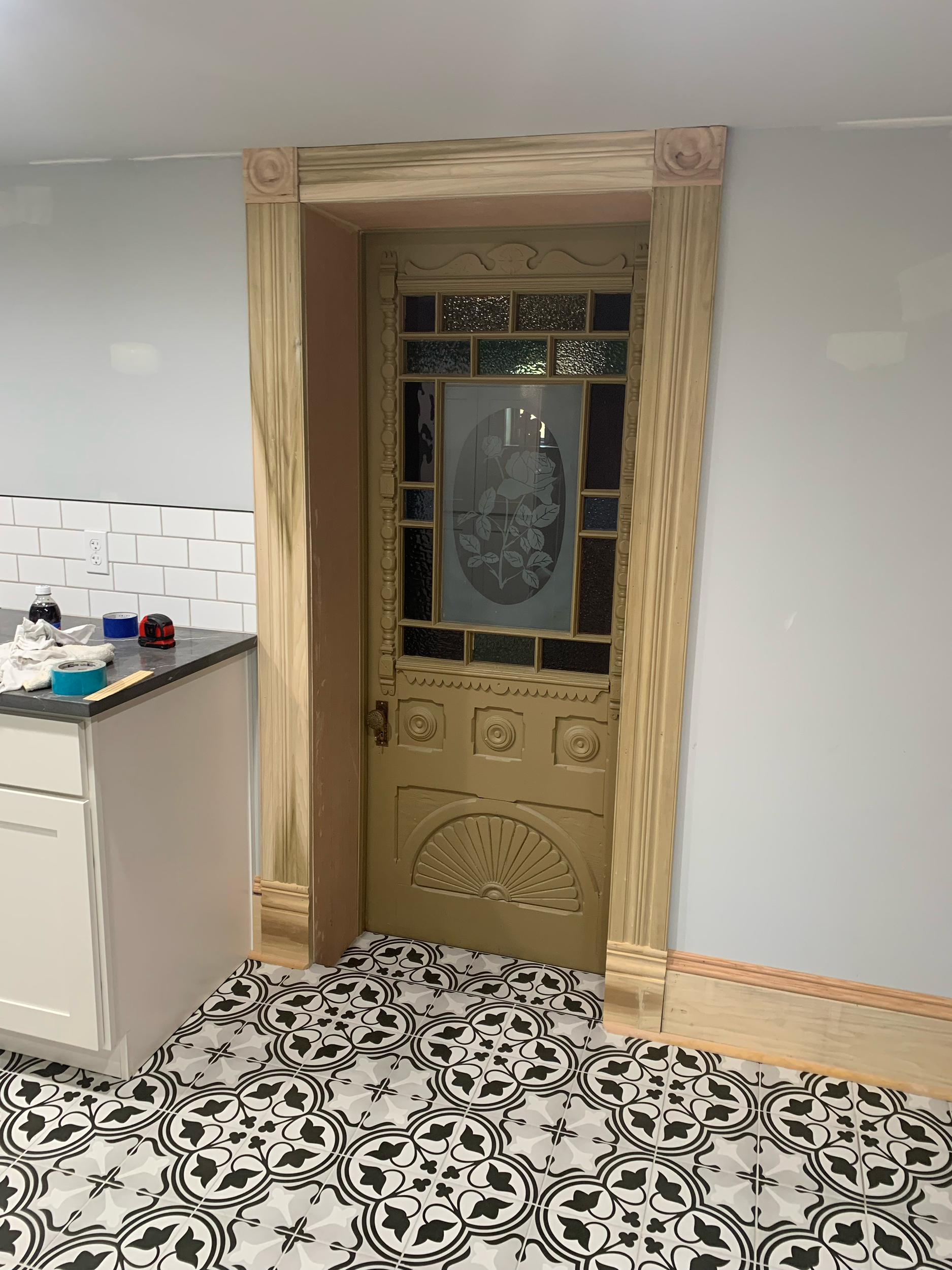 Decorative entrance door with glass panels and floral etched design, surrounded by light wood trim, in a room with patterned black and white ceramic floor tiles, white wall, countertops with tools and supplies.
