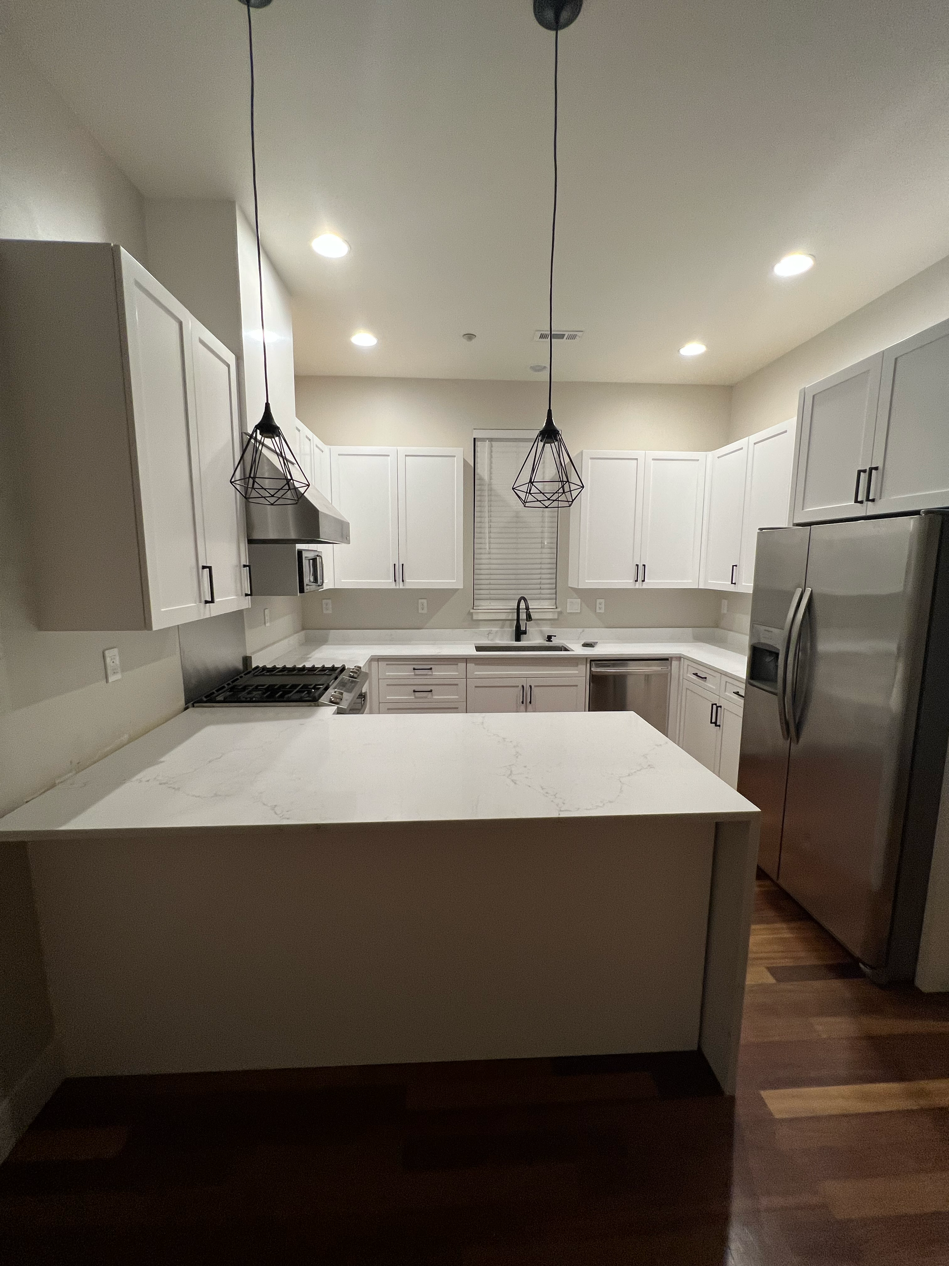 Modern kitchen with white cabinets, marble countertop island, stainless steel appliances, black pendant lights, and a window above the sink.