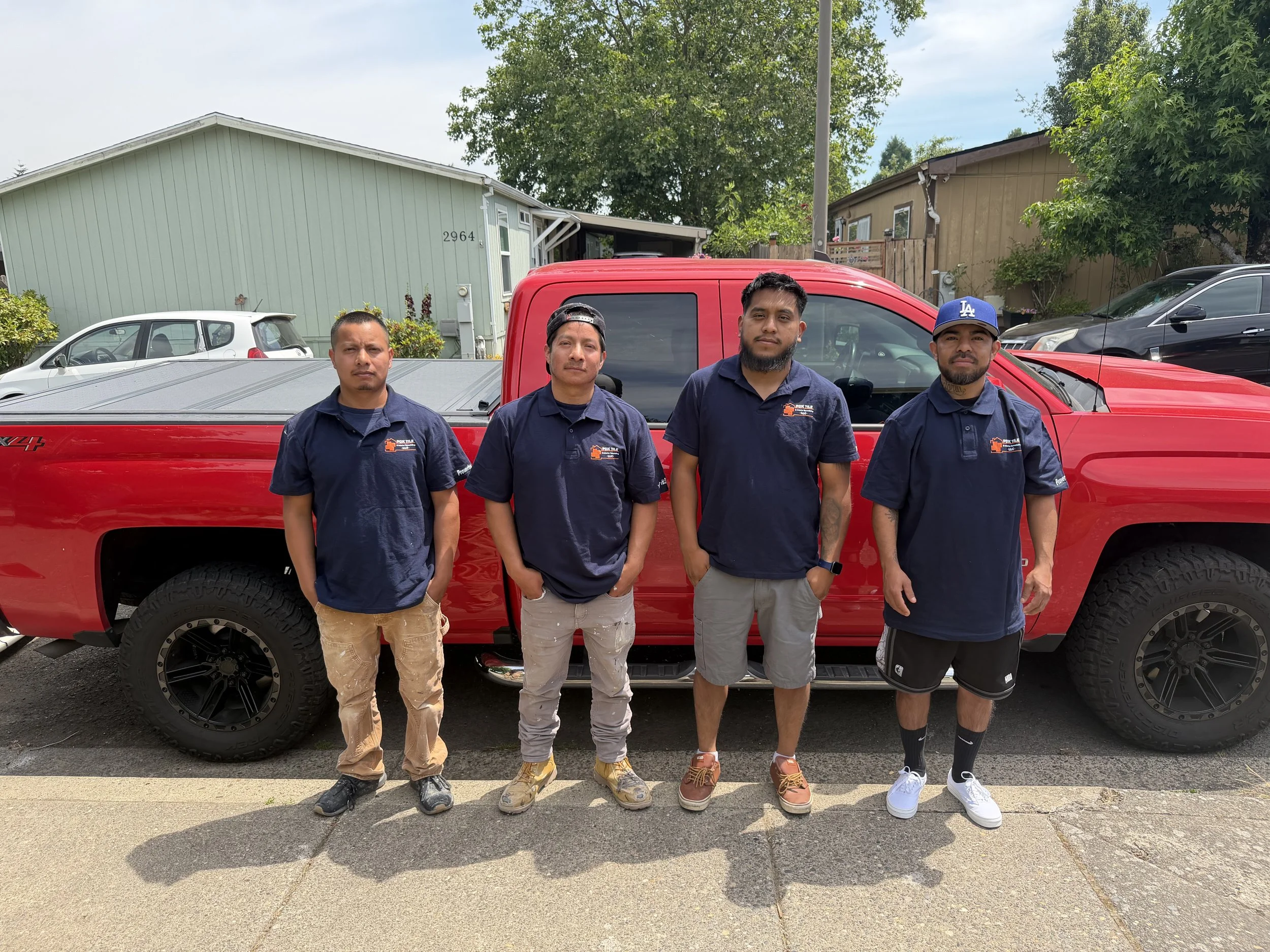 Four men standing in front of a red pickup truck on a residential street, with houses and trees in the background.