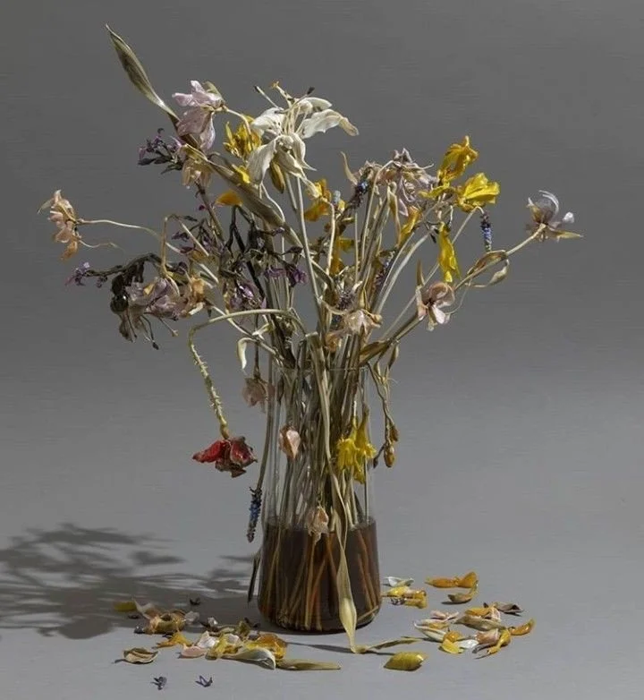 A glass vase with wilted and dried flowers, with some petals scattered around the base, set against a plain gray background.