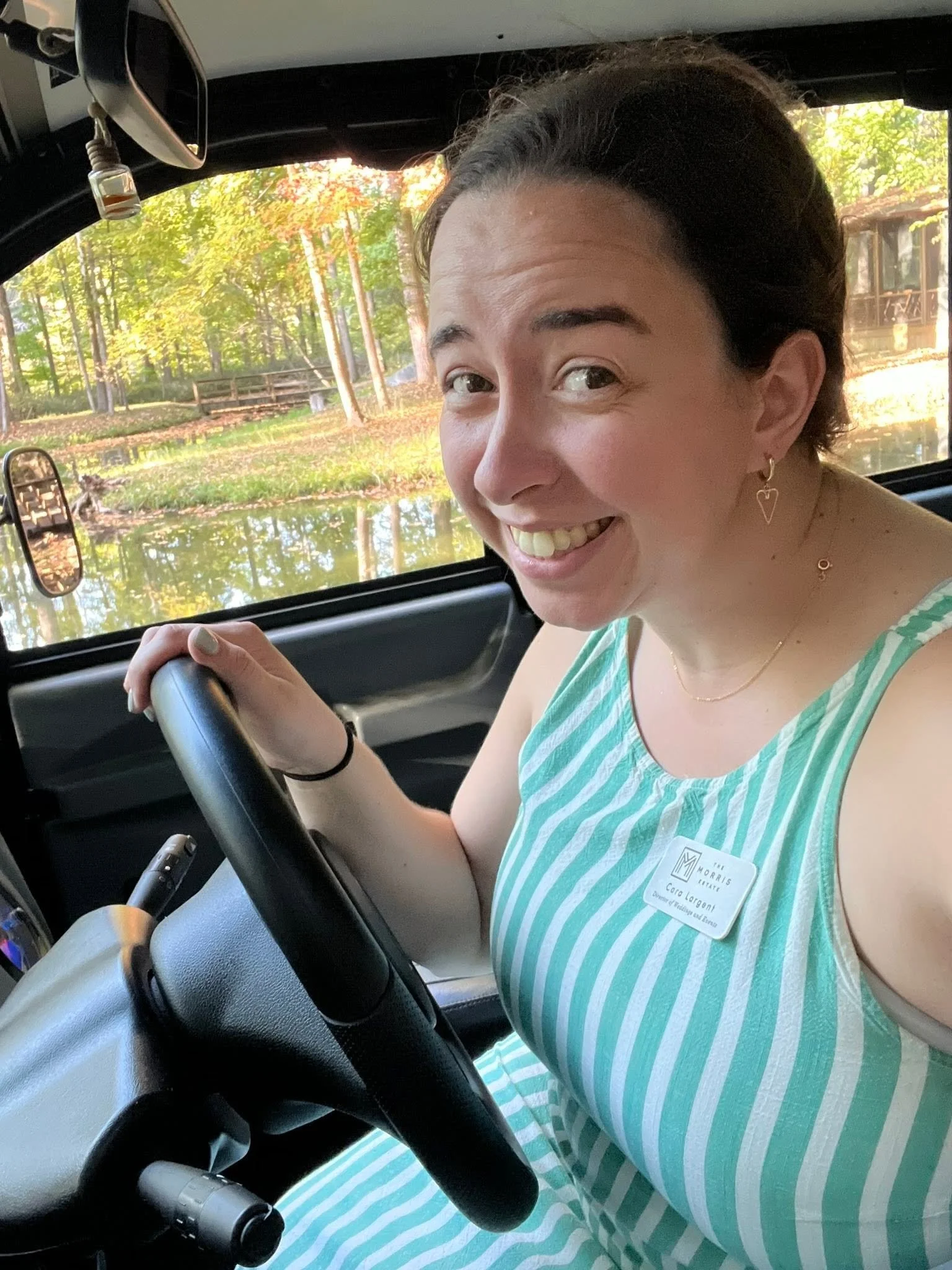 A woman with dark hair smiling while sitting in the driver's seat of a vehicle, holding the steering wheel. She is wearing a striped sleeveless top and a name tag.