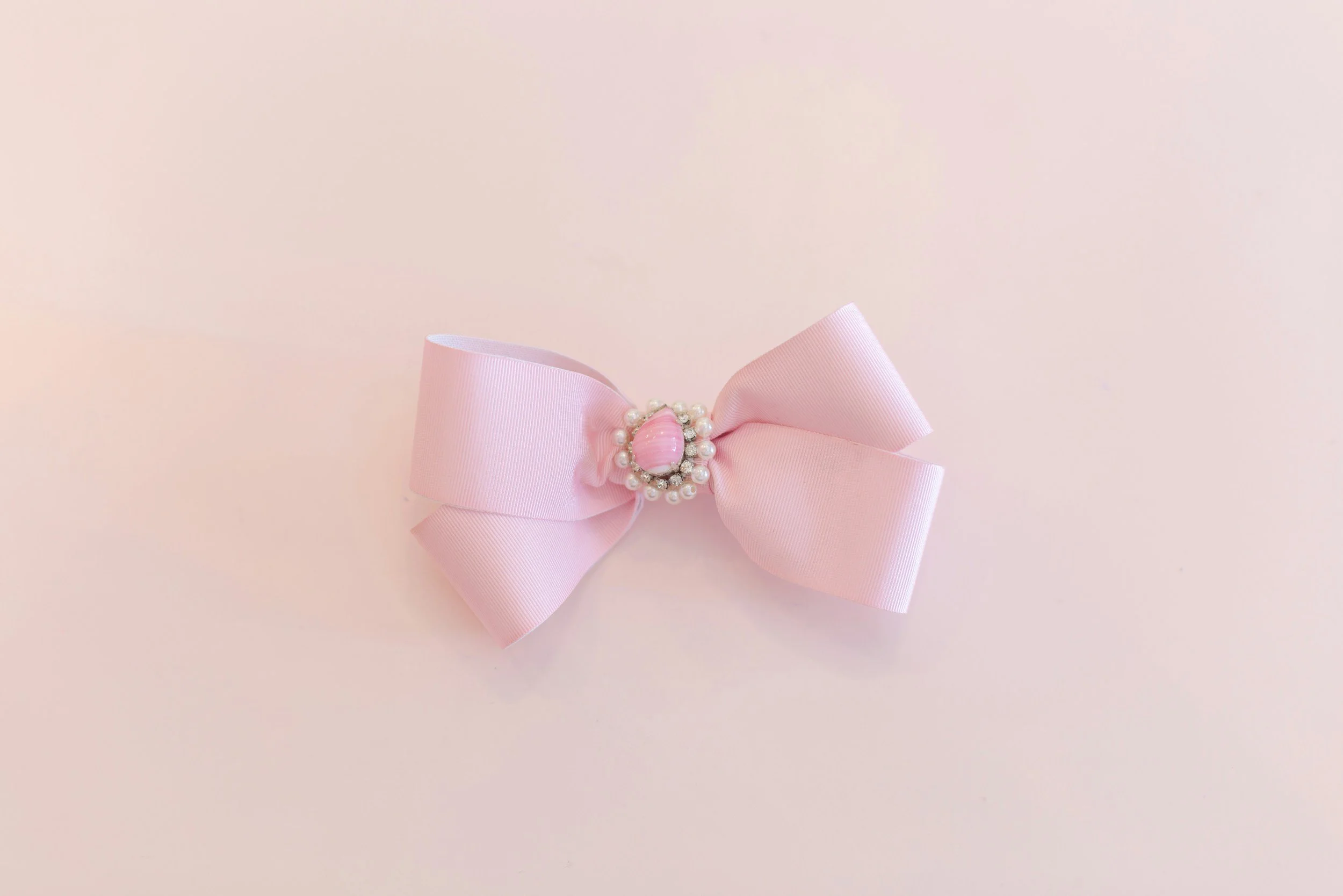 A pink ribbon bow with a central decorative piece made of pearls and a pink gemstone, placed on a light pink background.