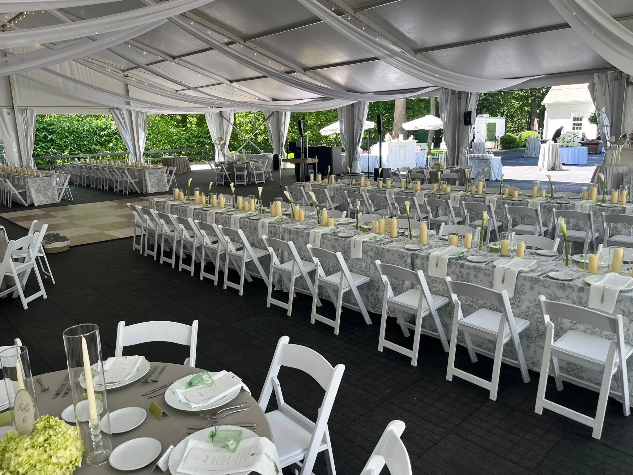 An indoor wedding reception area with white drapes and bistro lights on the ceiling. Round and long rectangular tables are decorated with floral tablecloths, candles, and white flowers. Greenery is visible outside through open sides.