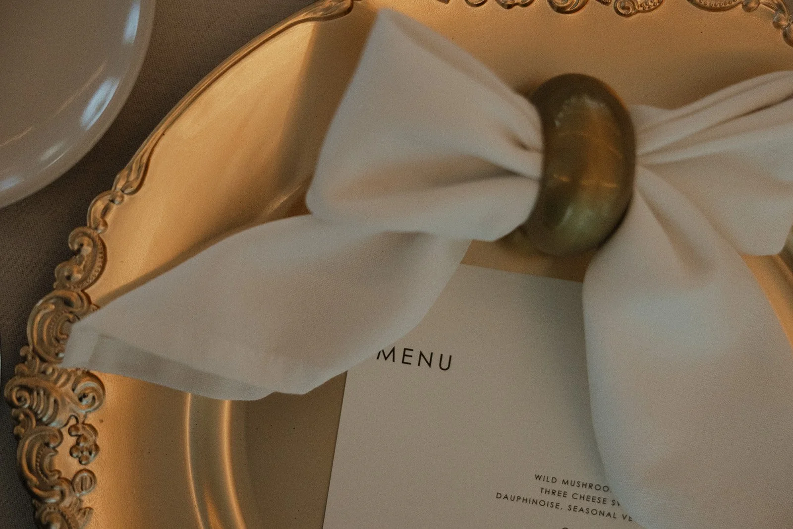A decorative gold charger holding a white napkin, a menu, and a white plate in the background. The tray has ornate edges and a gold napkin ring holder with a white napkin. The menu includes a dish with wild mushroom, cheese, and dauphinoise.