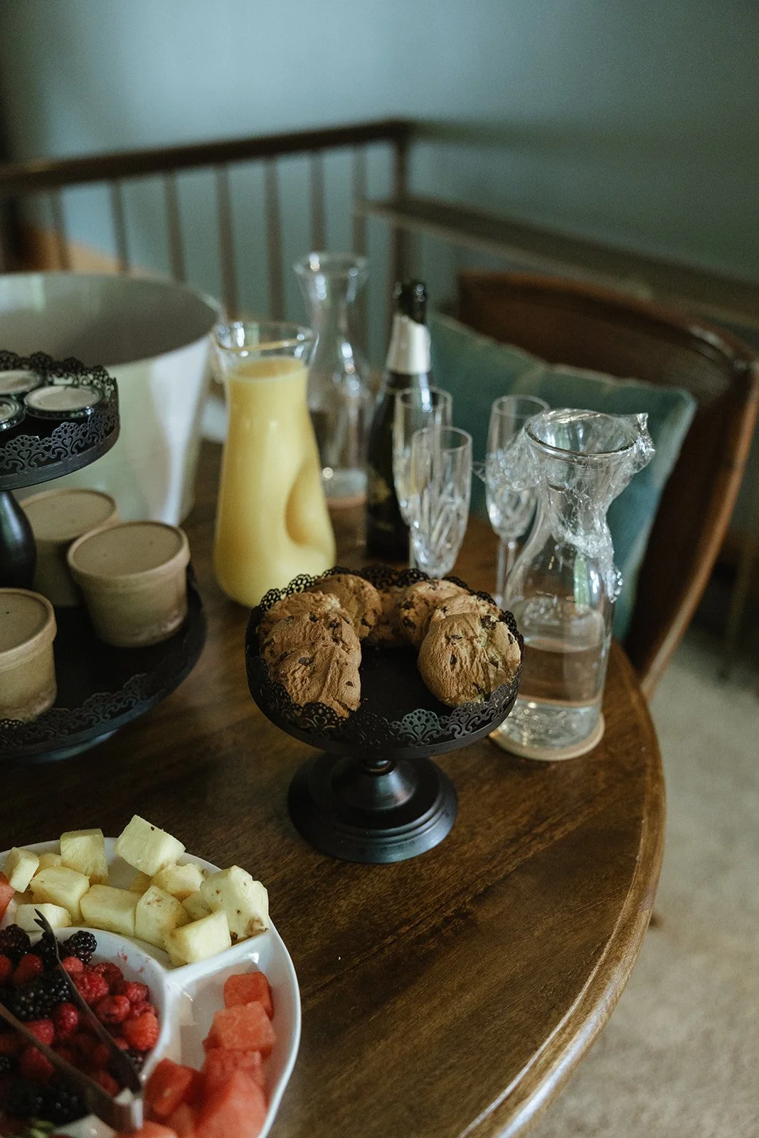 A table set with glass pitchers, champagne flutes, a yellow pitcher, candles, a plate of cookies, and a fruit platter.