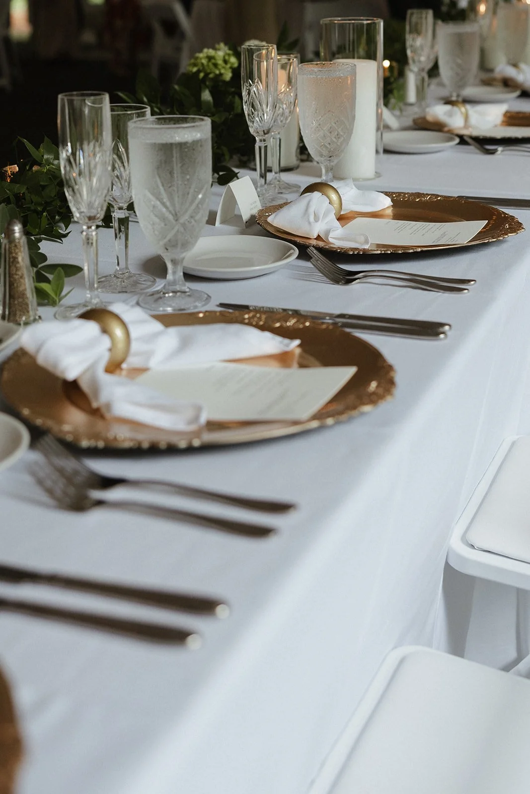 Elegant table setting with gold chargers, white napkins, clear glassware, and greenery decoration for a formal event or dinner party.