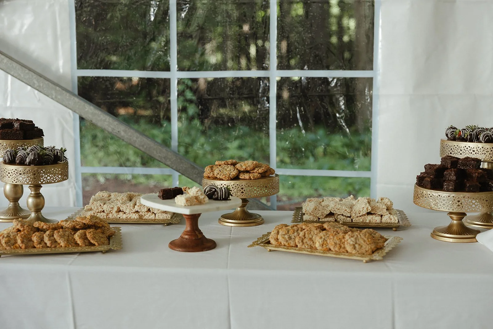 Dessert table with various cookies and chocolates displayed on gold stands, set against a window view of greenery.