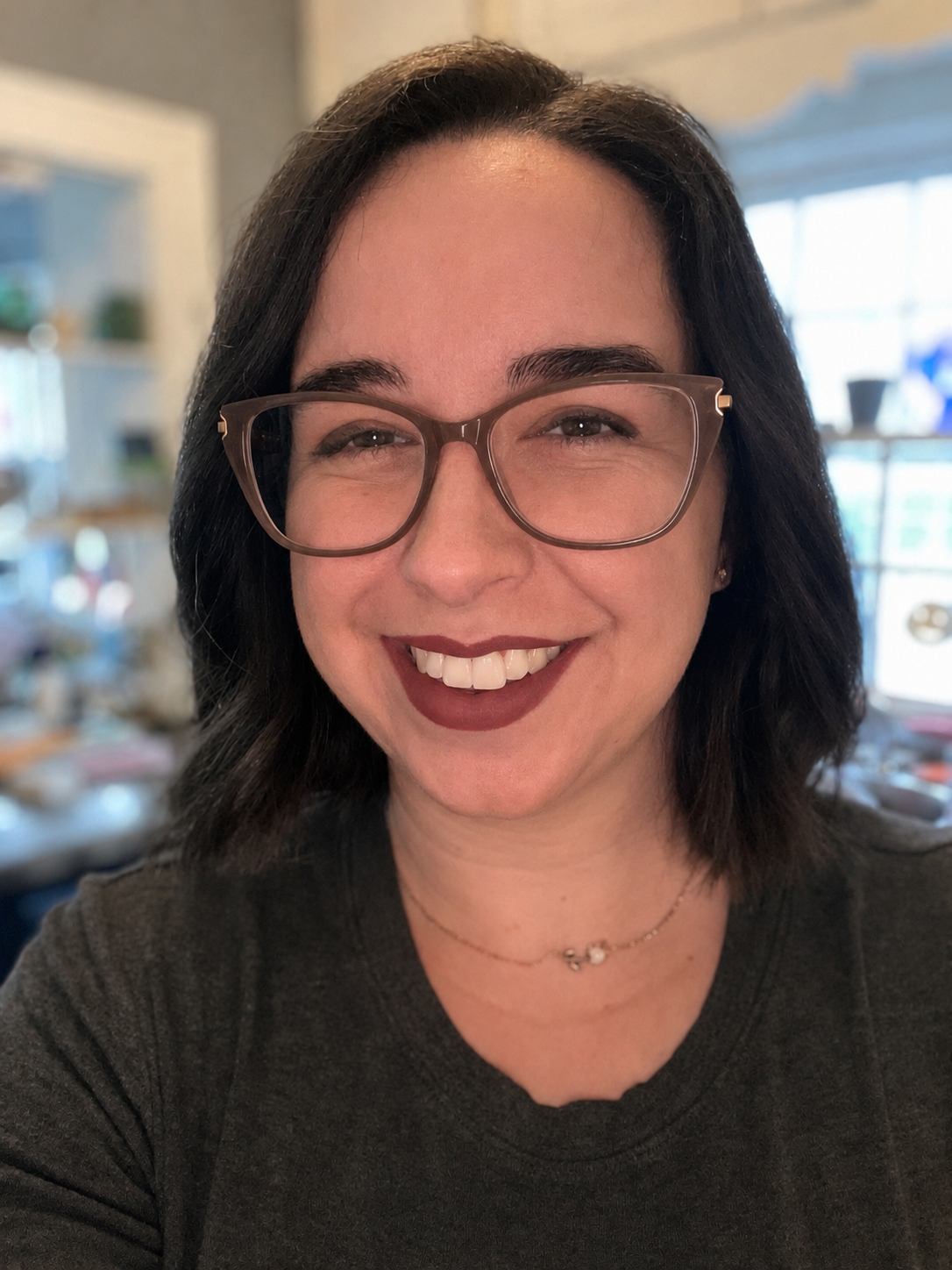 Close-up of a woman smiling with glasses and dark wavy hair, in a cozy indoor setting with bookshelves and windows in the background.