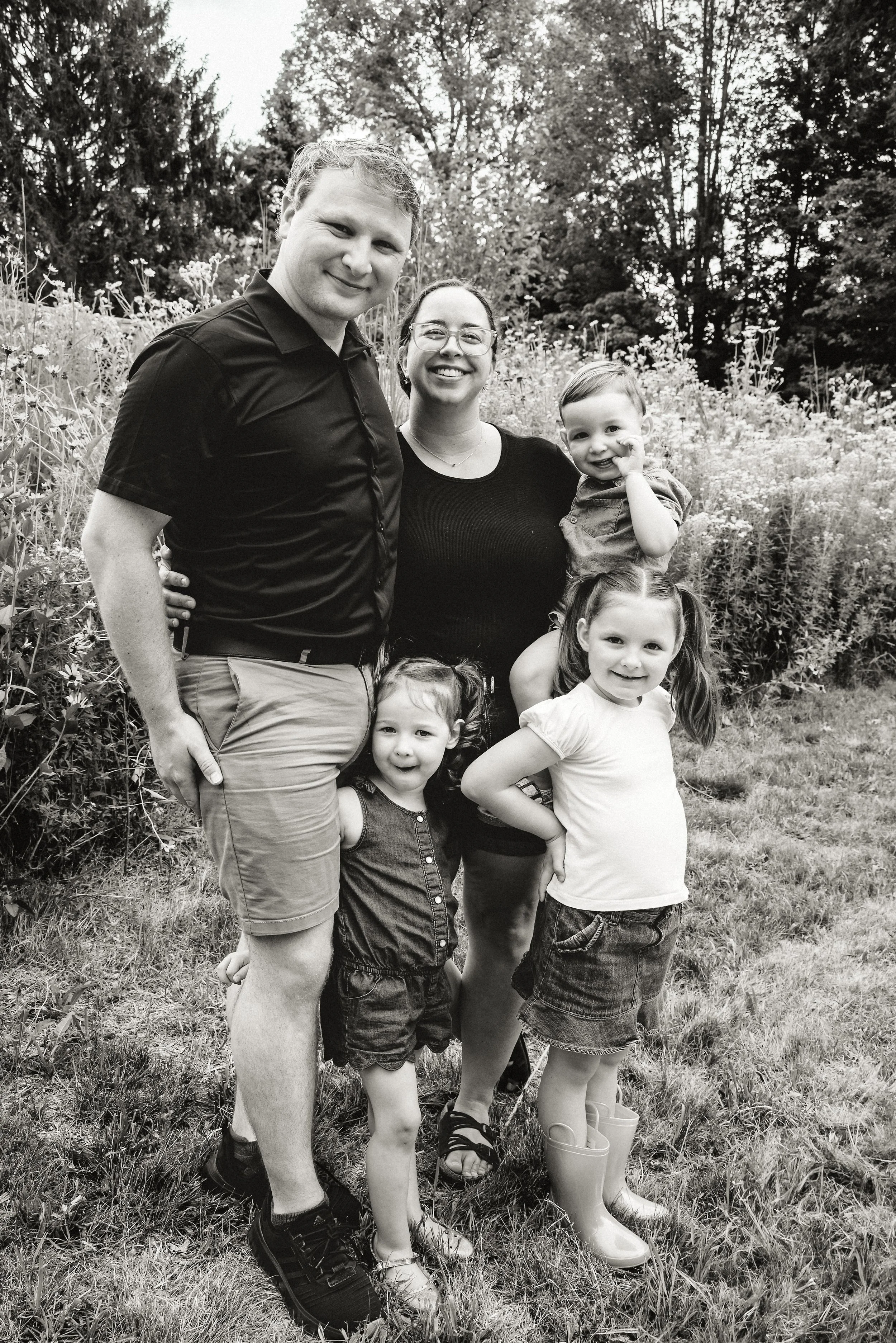 A black and white photo of a family of five standing outdoors in a garden with trees and flowers in the background, smiling at the camera.