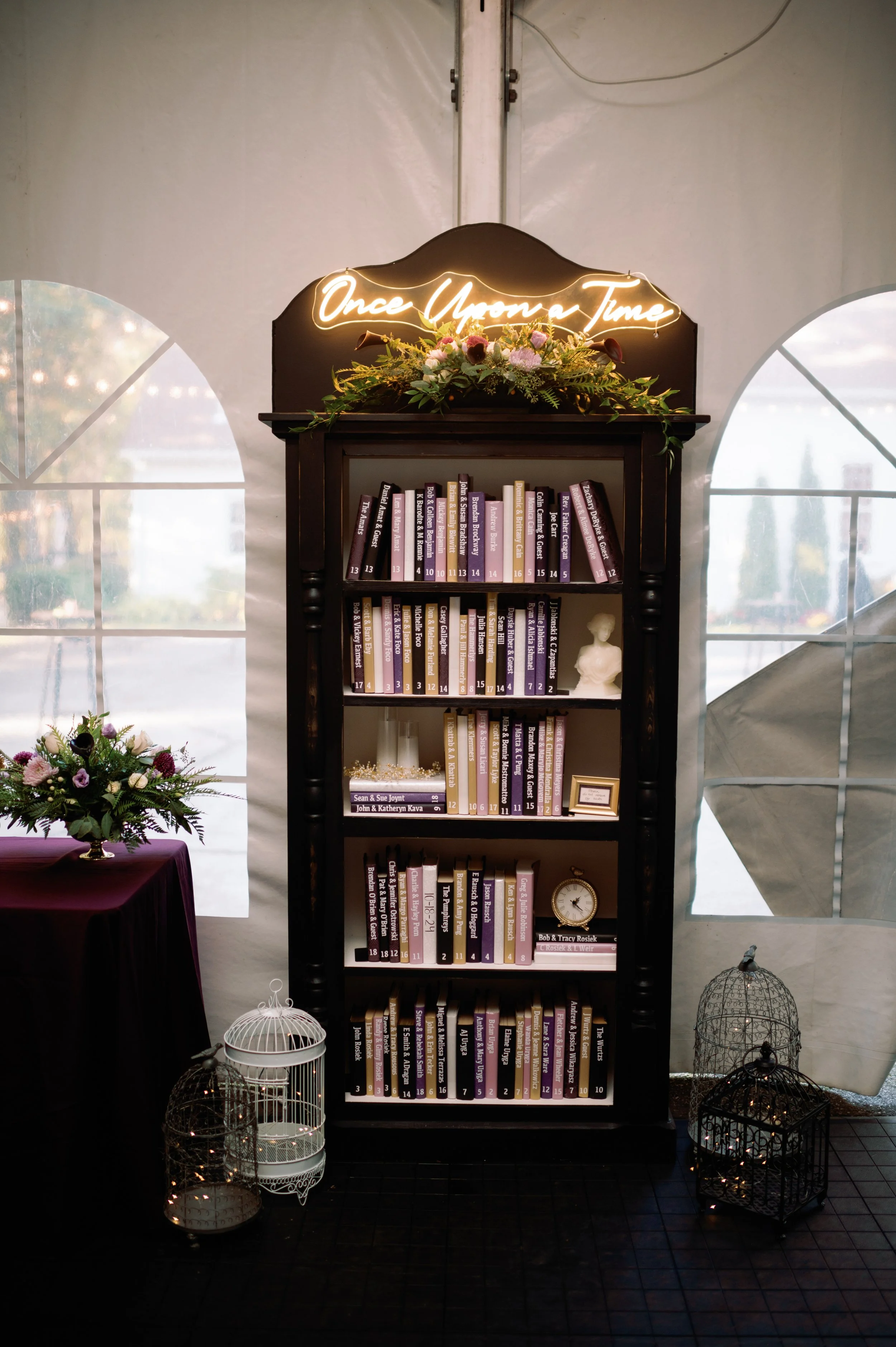 A black bookshelf with five shelves filled with books, decorated with a floral arrangement on top and a neon sign that reads 'Once Upon a Time'. To the left, a table with a purple cloth and a floral centerpiece, and to the right, decorative birdcages on the floor. The background features white fabric walls with windows.