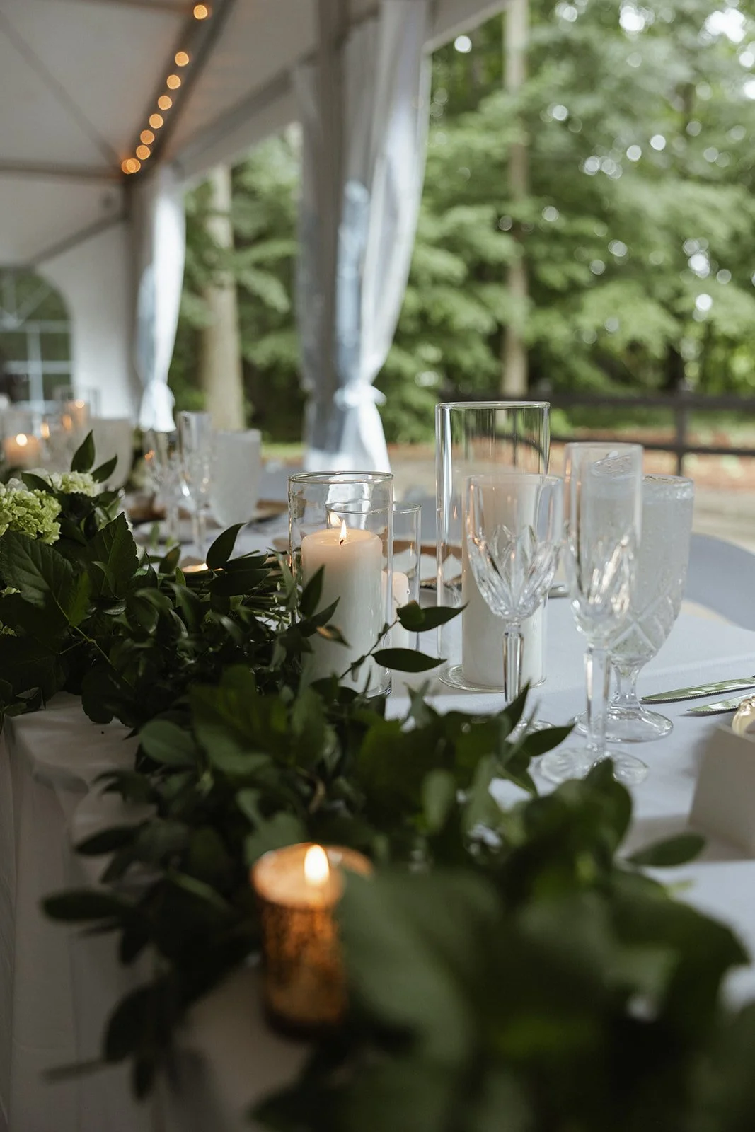 Wedding reception table decorated with candles, greenery, and elegant glassware inside a tent with a view of trees outside.