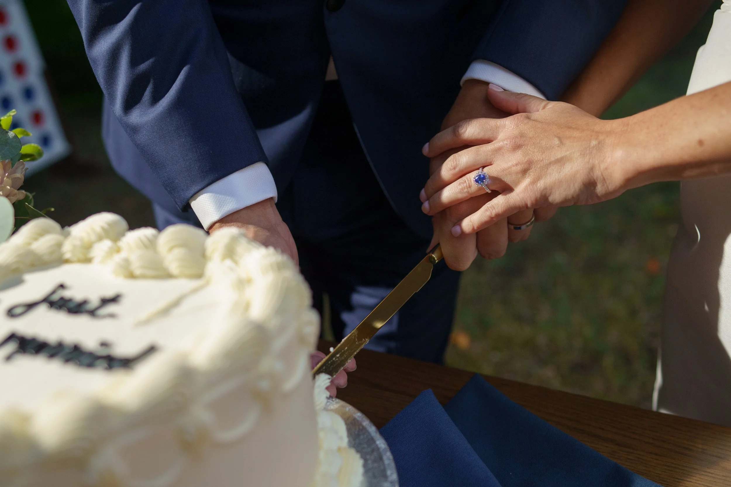 A couple is cutting a wedding cake together, holding a knife. The man is wearing a dark suit with a white shirt, and the woman has a ring on her finger. The cake is decorated with white frosting and has the word 'love' written on it.